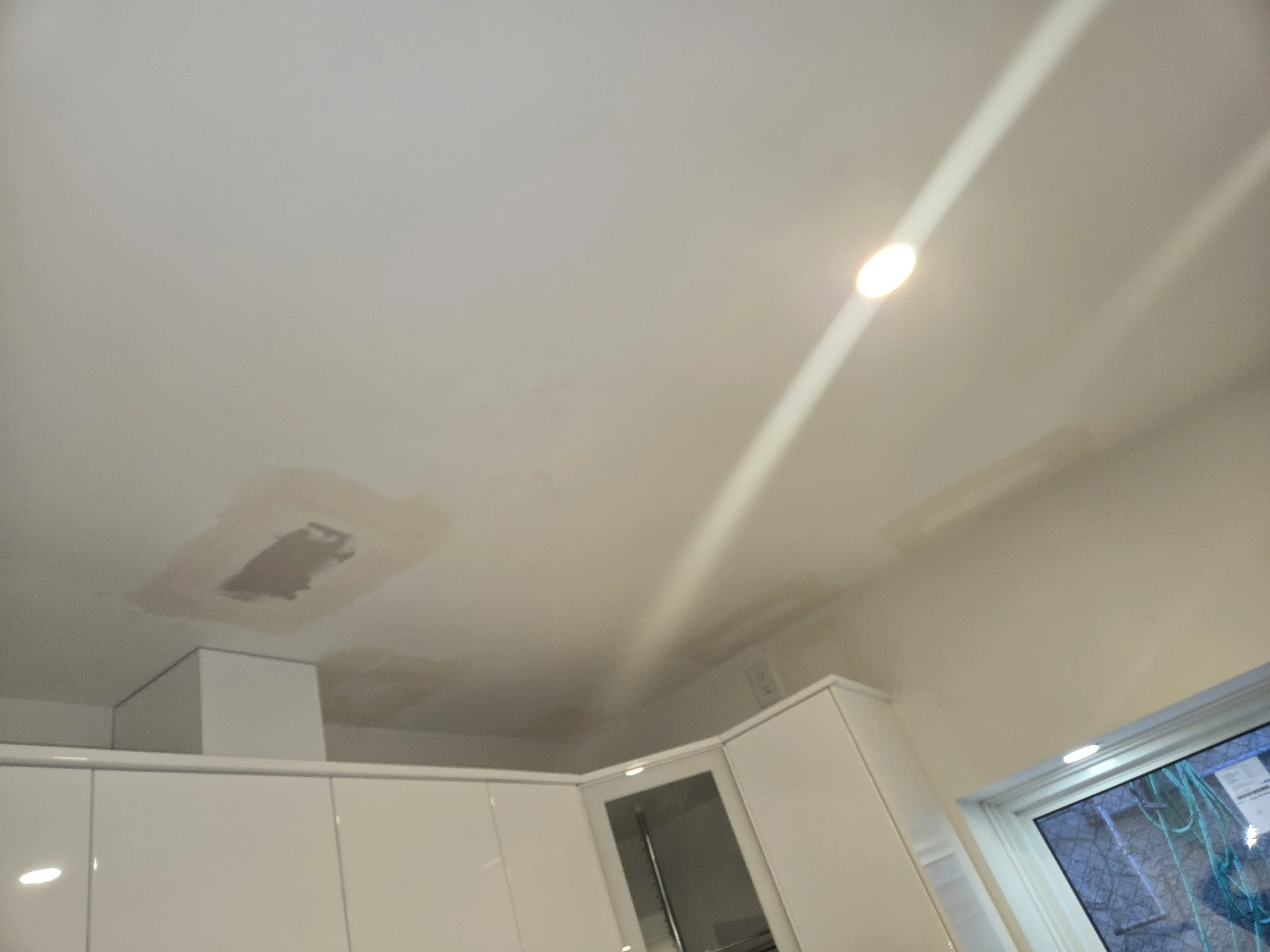 Ceiling with patches of dried paint or patching compound and a bright ceiling light, part of a kitchen with white cabinetry and a window.