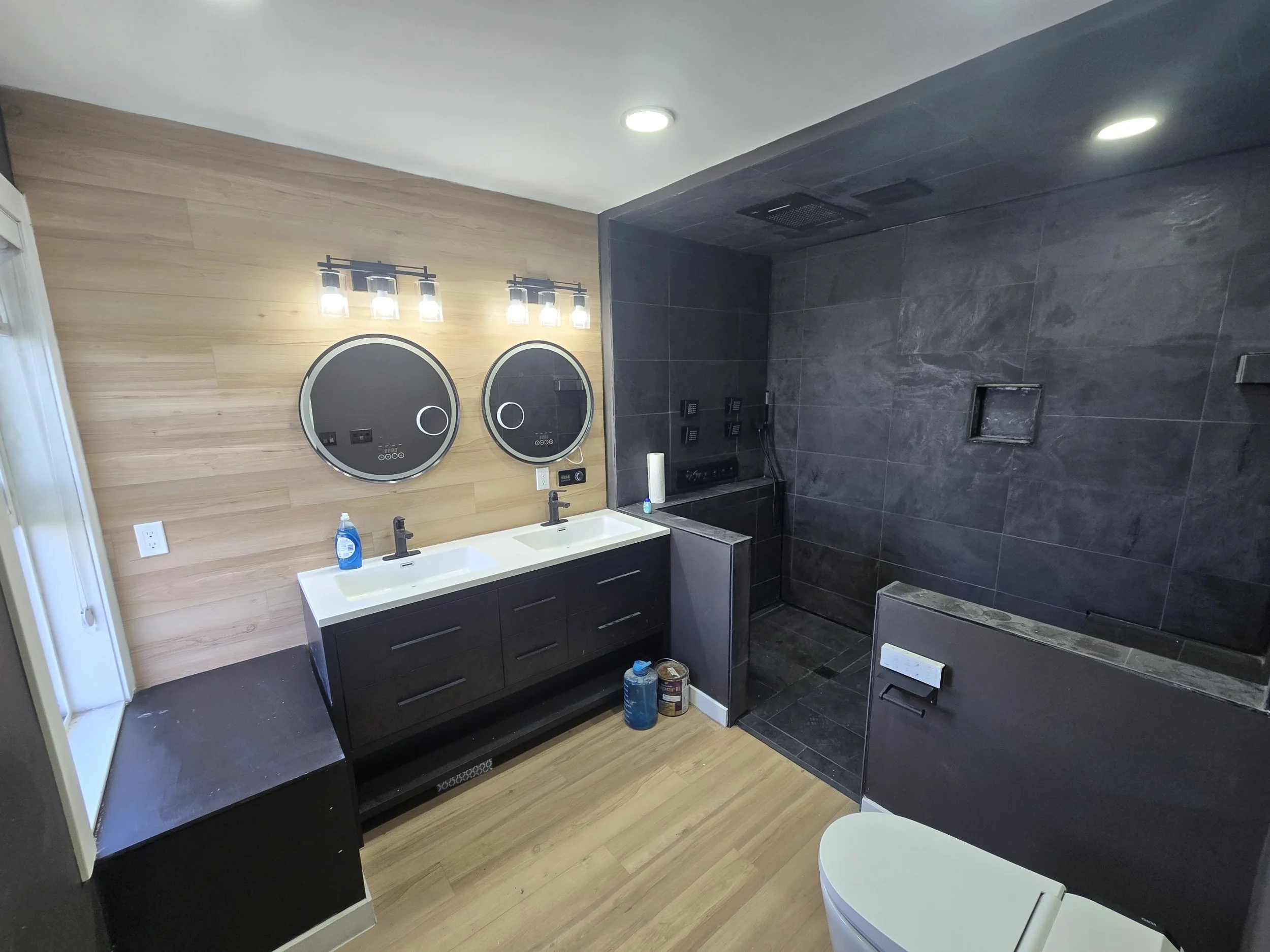 Modern bathroom with dual sinks, circular mirrors, black cabinetry, wooden wall paneling, black tile shower area, and wooden flooring.