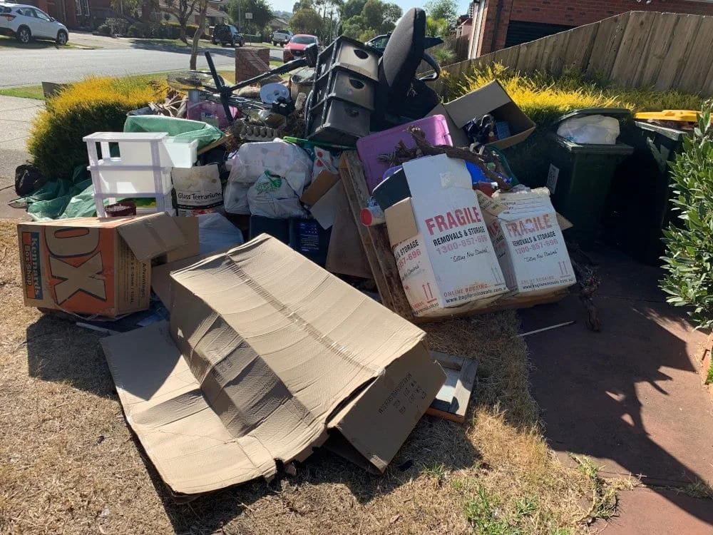 Piles of garbage and discarded items, including cardboard boxes, plastic storage bins, and household clutter, outside on the grass next to a sidewalk and driveway.