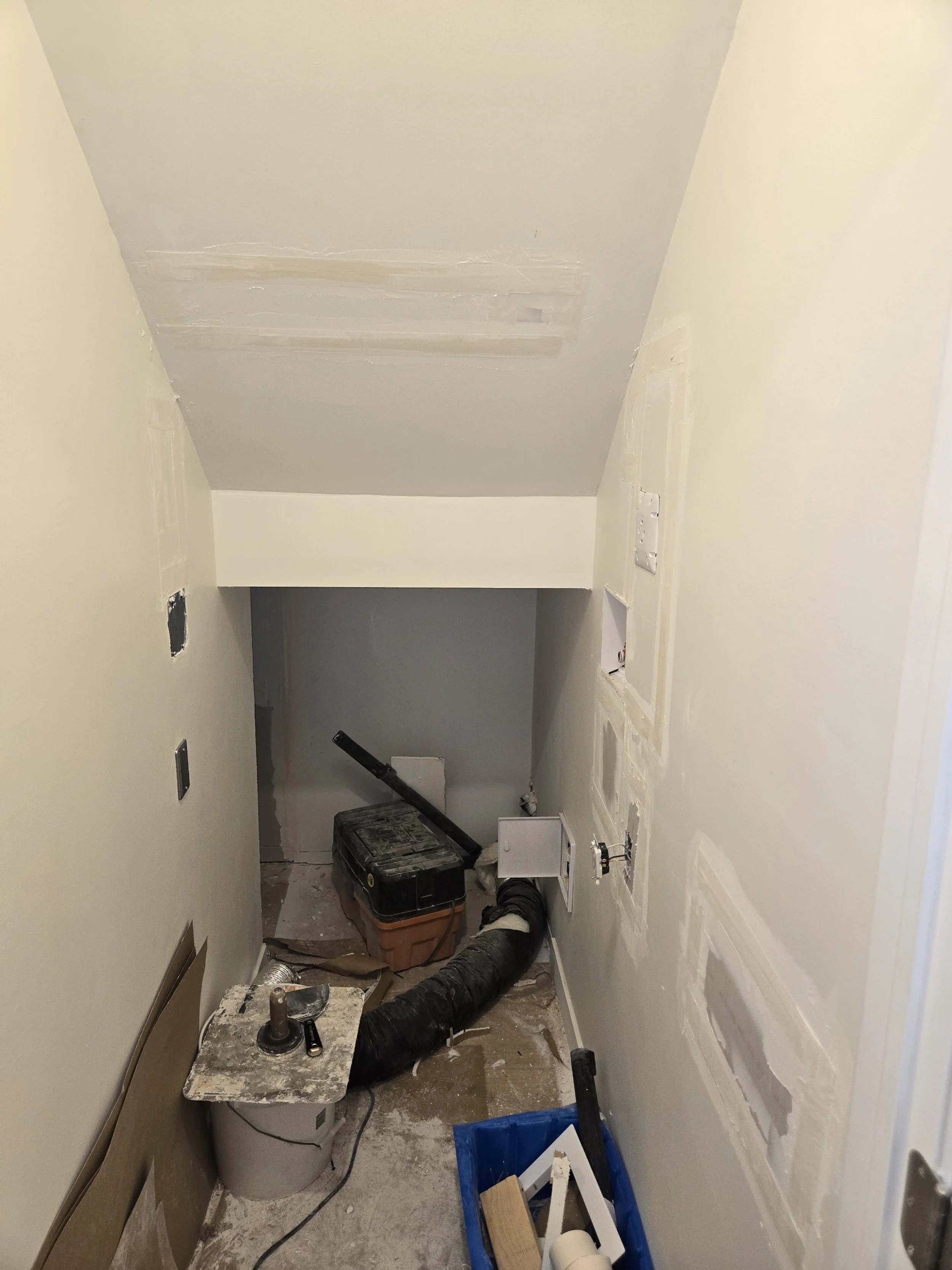 Small, narrow utility room under renovation with exposed drywall and construction tools, including a toolbox, power drill, ducts, and electrical outlets.