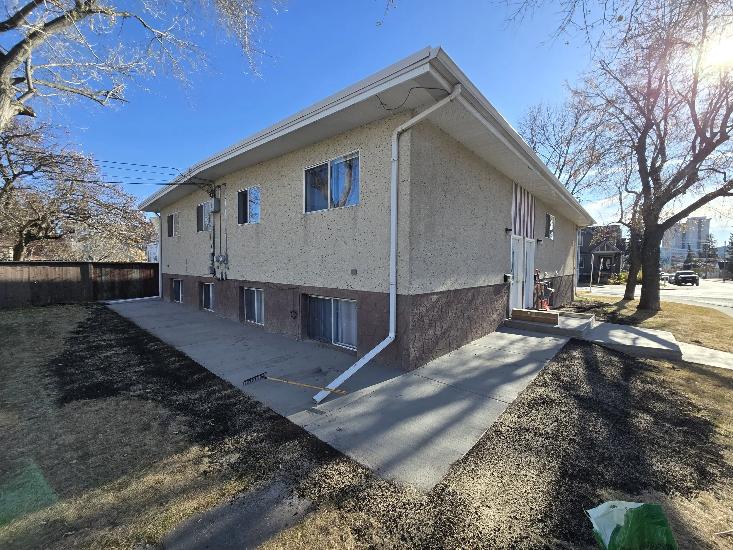 Side view of a beige two-story house with a concrete walkway, leafless trees, and a clear blue sky.