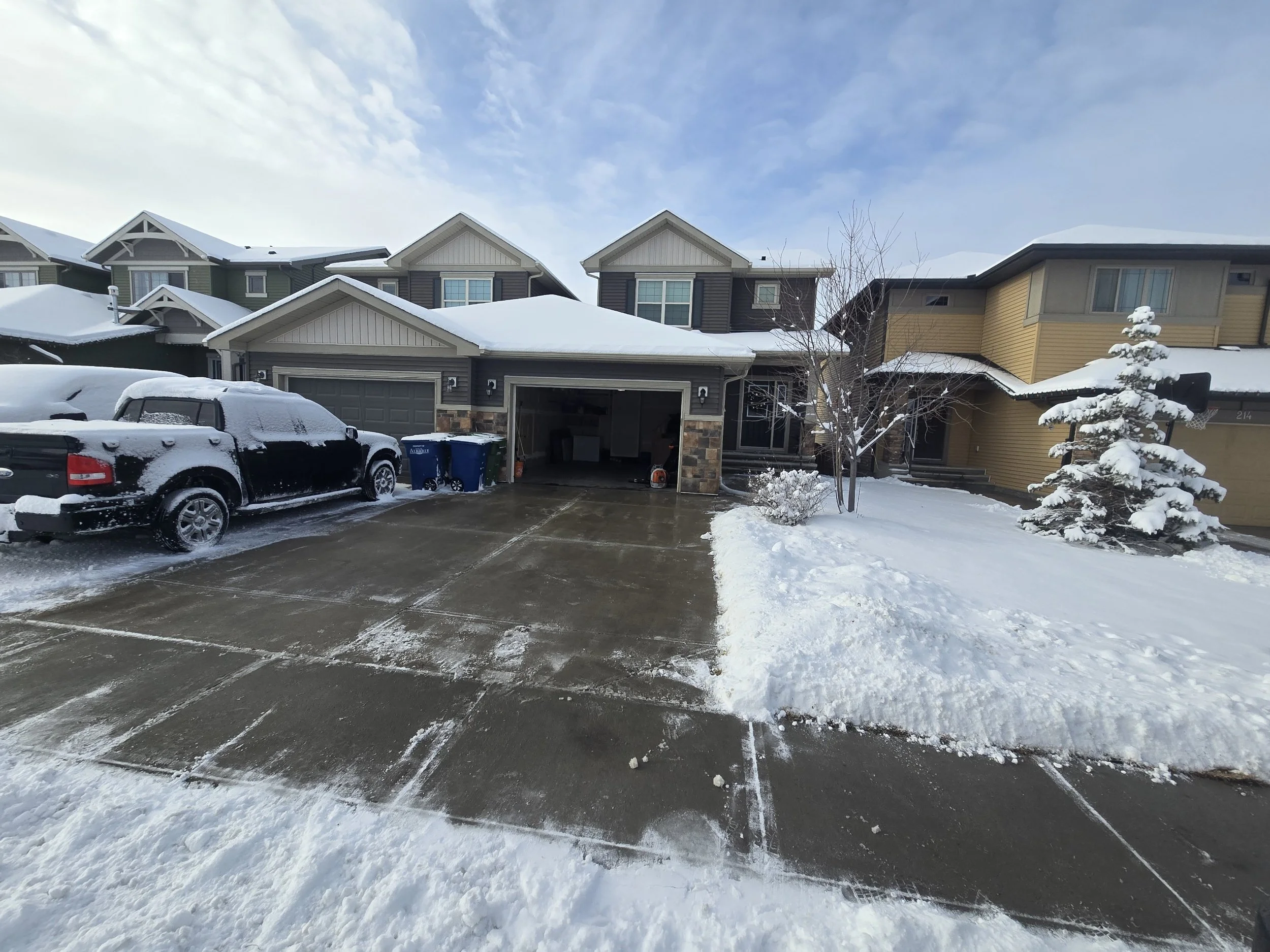 A snow-covered suburban house with a driveway, parked SUV, and blue trash bins. Snow is on the roof, trees, and ground, and the sky is partly cloudy.