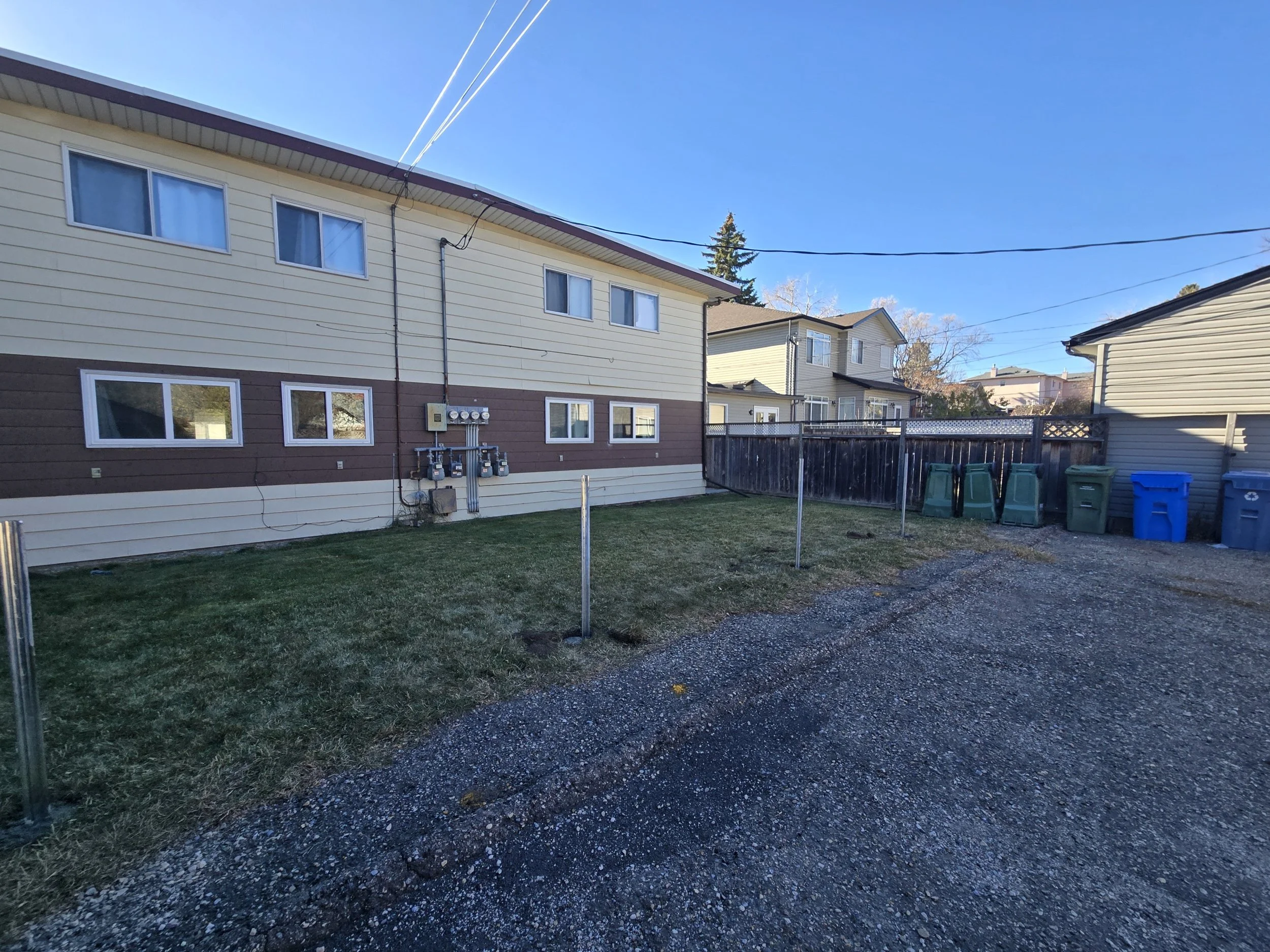 Backyard with green grass, gravel driveway, house with beige siding and brown accents, utility meters, and multiple garbage and recycling bins near a wooden fence under a clear blue sky.