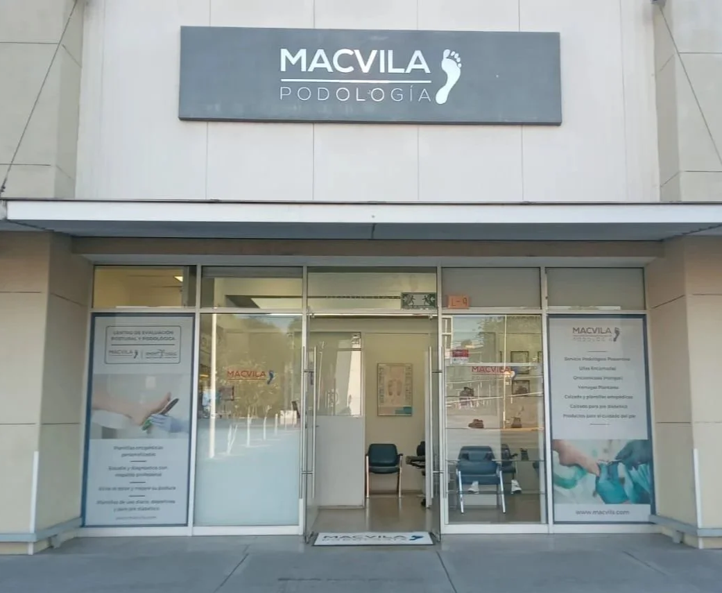 Entrada de una clínica de podología llamada MACVILA, con ventanas de vidrio y una señal grande en la parte superior con el logotipo de un pie, indicando que es un servicio de podología.
