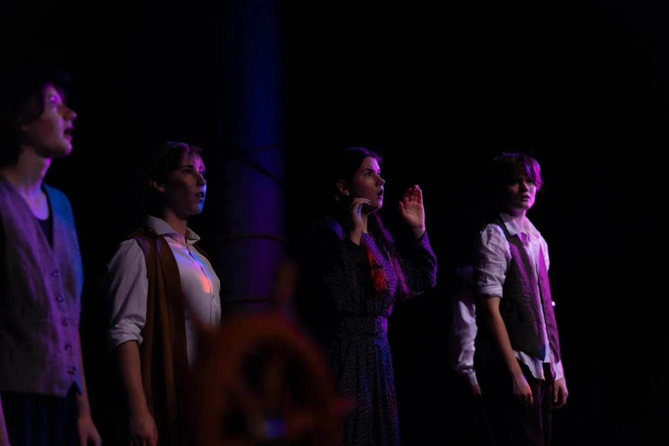 Four actors are bathed in dark pink light. Three are wearing vests and shirts, and one has two long ponytails and a purple dress. All of them look shocked. There is a blurry ship's wheel in the foreground.
