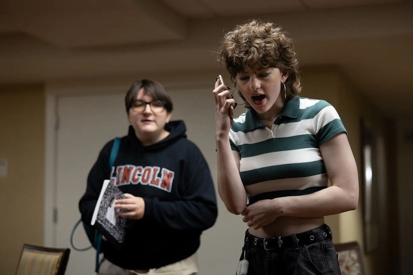An actress in a striped white and green shirt is yelling into a cellphone. Behind her, an actor in a sweatshirt holds a notebook.