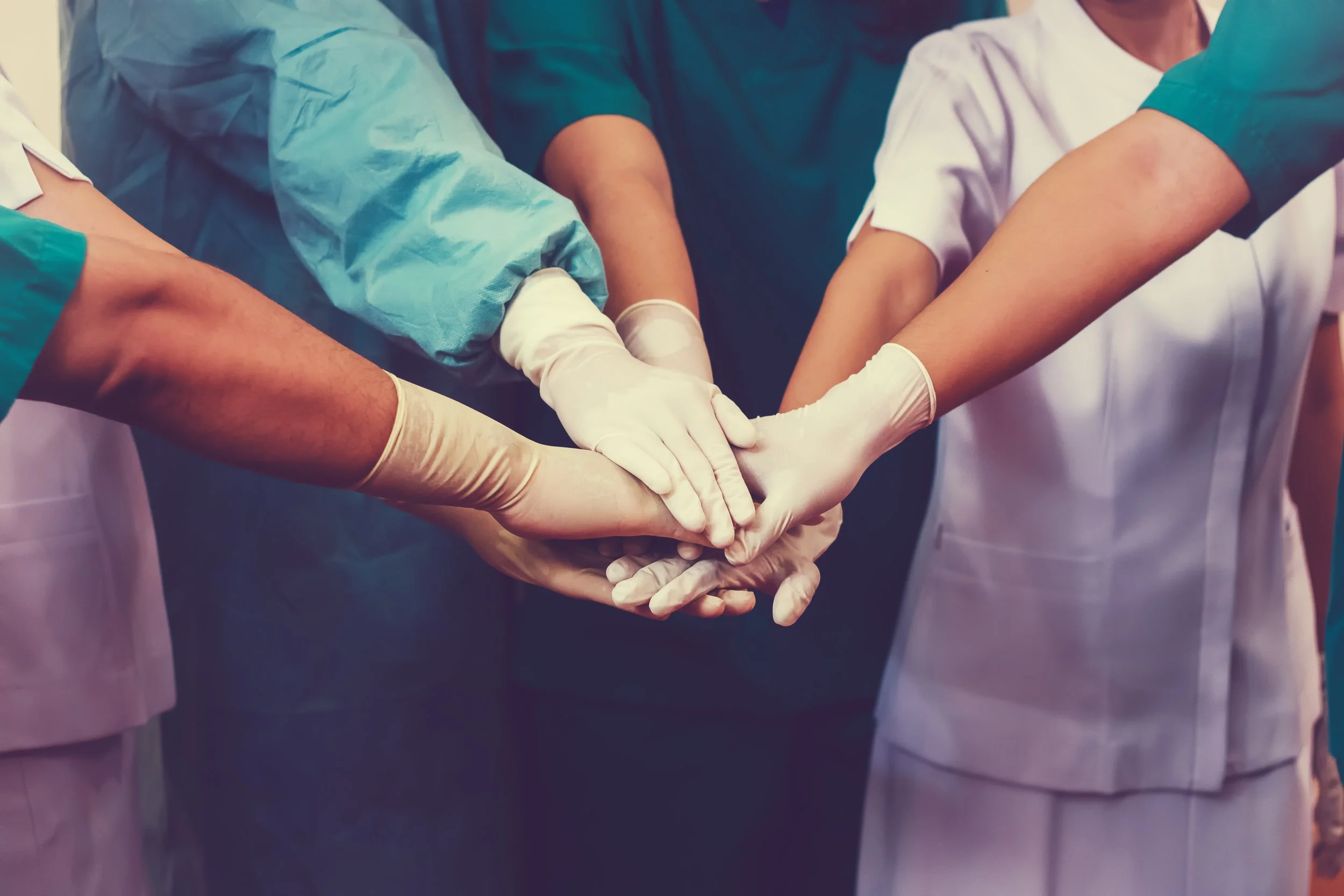 Group of healthcare professionals in scrubs and gloves putting their hands together in a team huddle.