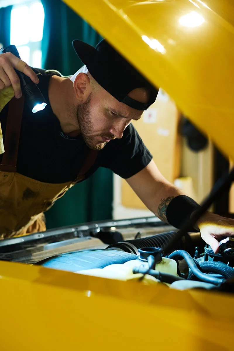 Auto mechanic in yellow overalls with flashlight working on yellow car