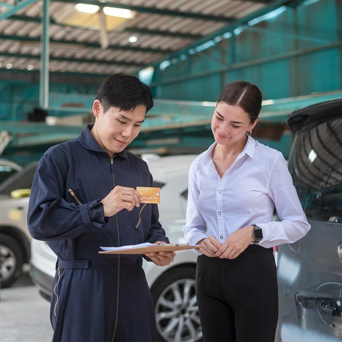 A man and a woman standing next to a car in an indoor garage. The man is holding a credit card and a clipboard, while the woman is smiling and looking at him. The background includes other vehicles and a metal roof structure.