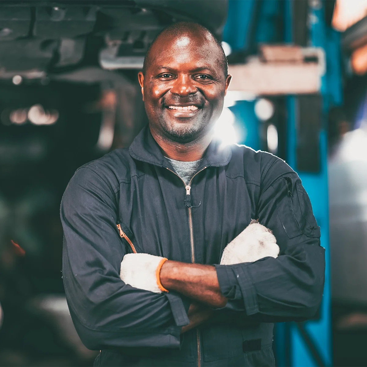 A smiling man in a mechanic's uniform standing in an auto repair shop with arms crossed.