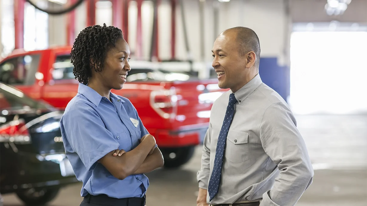 Female mechanic dressed in blue uniform, talking with a salesman in long sleeve khaki shirt and blue tie
