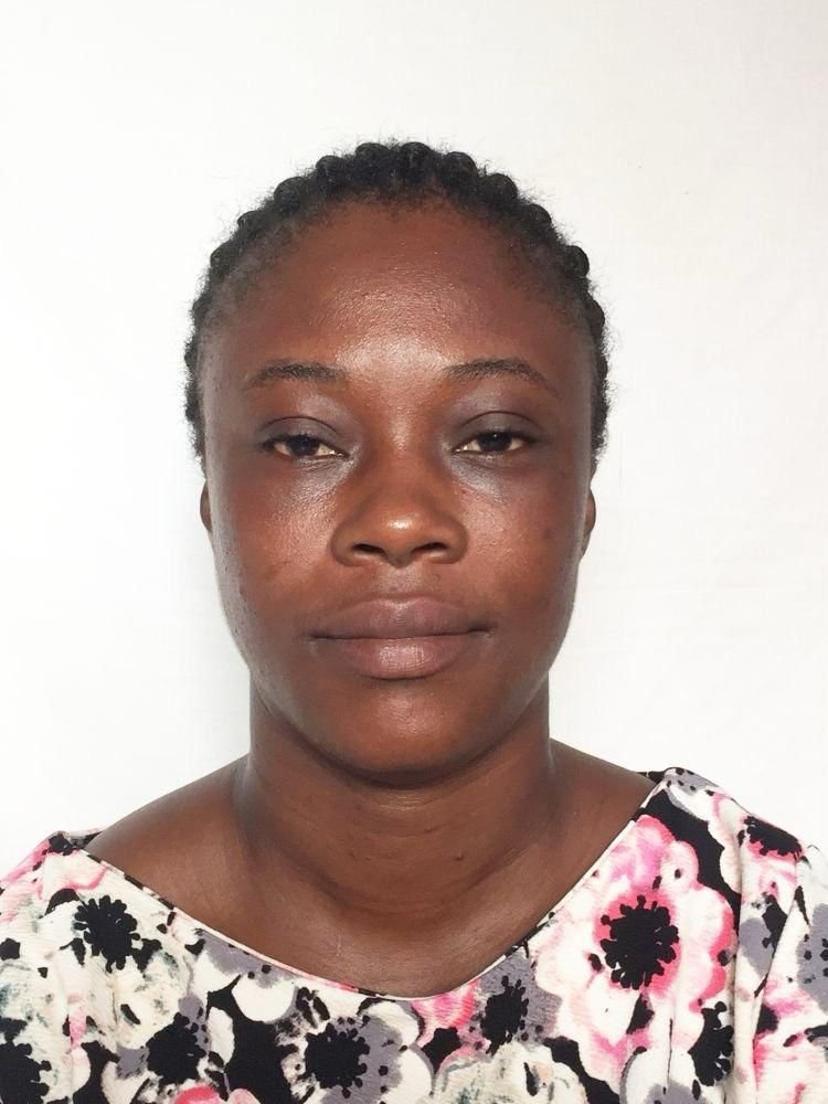 A woman with short, braided hair and medium brown skin, wearing a floral top, standing against a plain white wall.