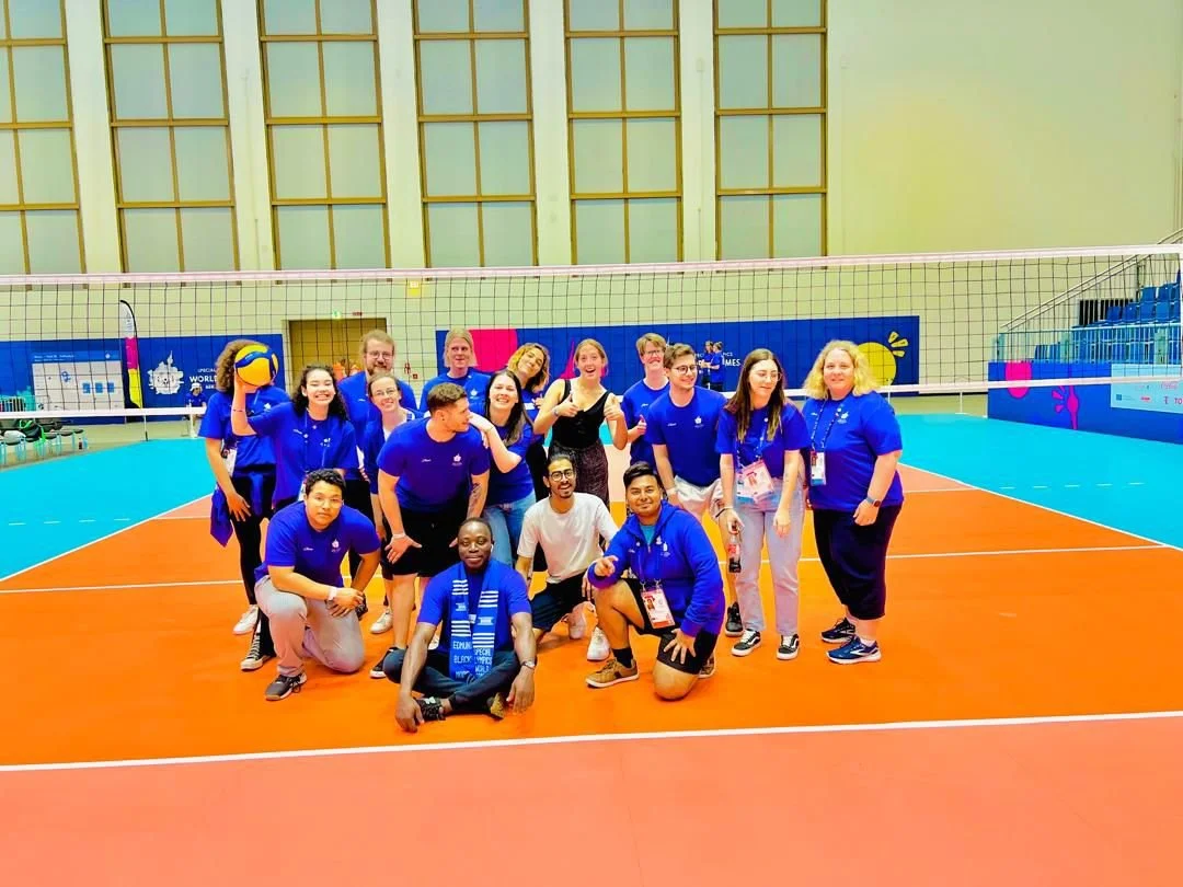 A group of people in blue shirts poses on an indoor volleyball court, some kneeling and some standing, with a volleyball net behind them.