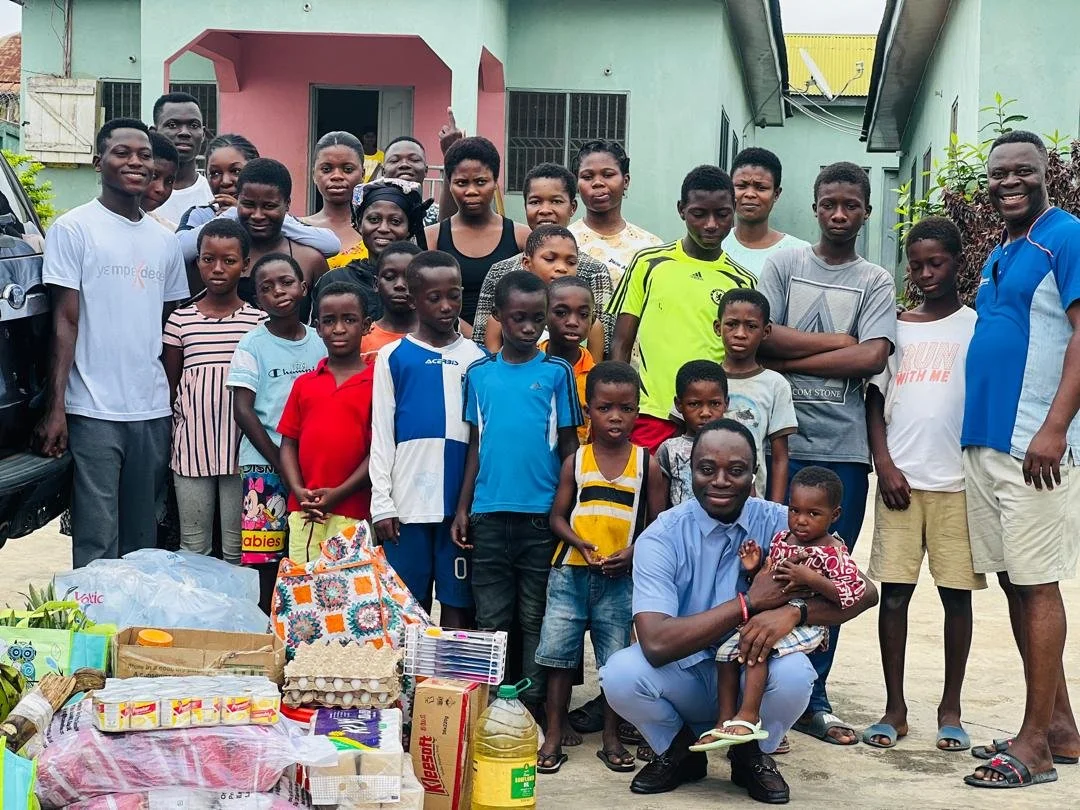 A group of children and adults posing outdoors in front of a building, with supplies and food items on the ground in front of them.