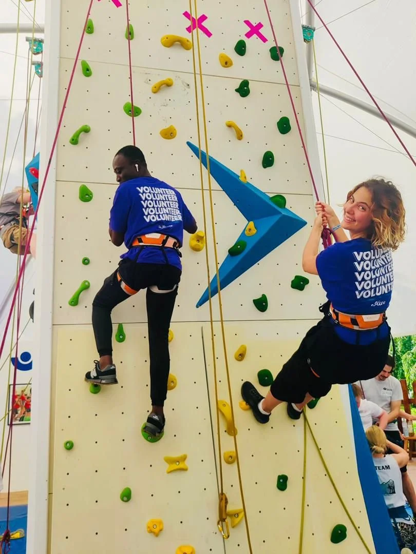 Two women climbing a rock wall at an indoor event, both wearing blue volunteer shirts and harnesses, with colorful handholds and footholds on the wall.
