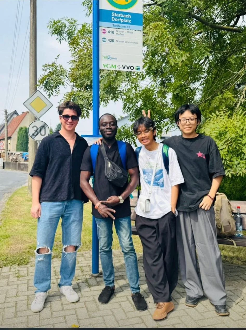 Four young people standing together at a bus stop in a residential area, smiling and posing for the photo. Behind them is a bus stop sign and some greenery.