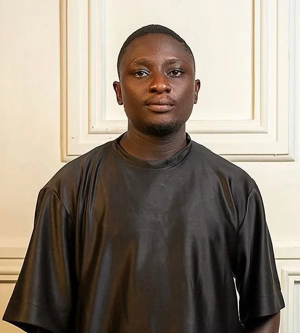 A young man with short hair and wearing a black shiny shirt standing in front of cream-colored paneled walls.