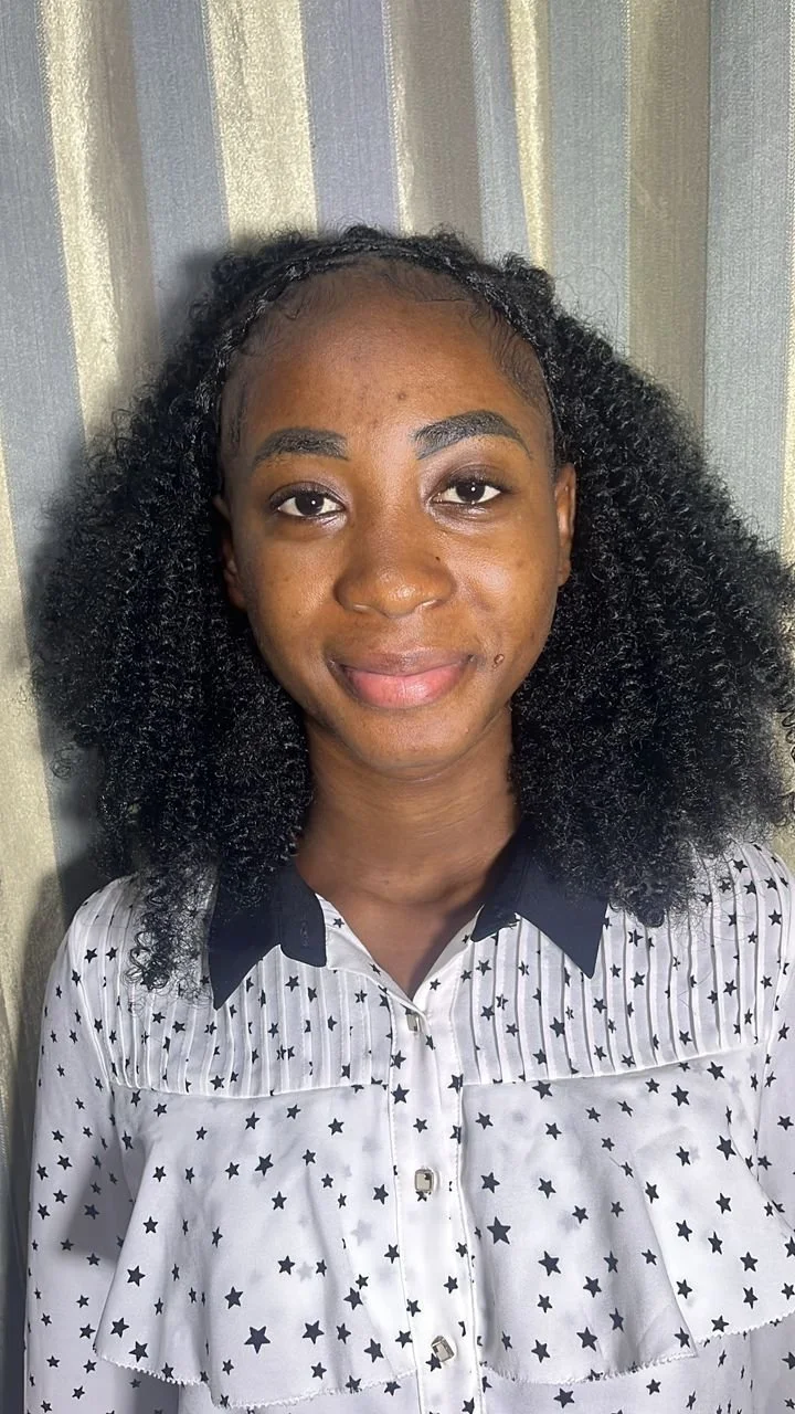 Close-up of a young woman with curly black hair, wearing a white shirt with black star patterns and a black collar, standing in front of a gray and beige striped curtain.
