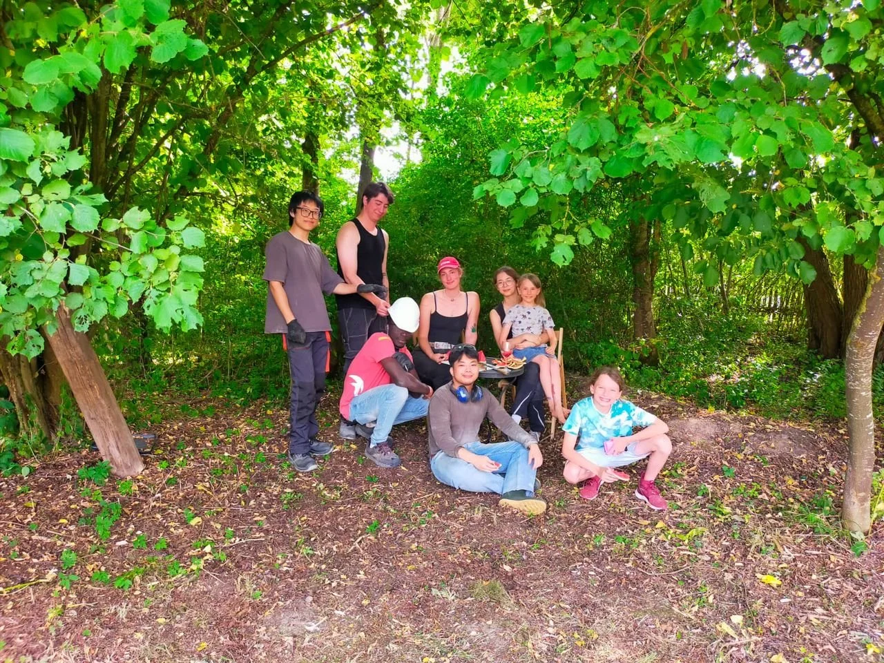 Group of people in a forest, some sitting on a log, others standing, with lush green trees and foliage surrounding them.