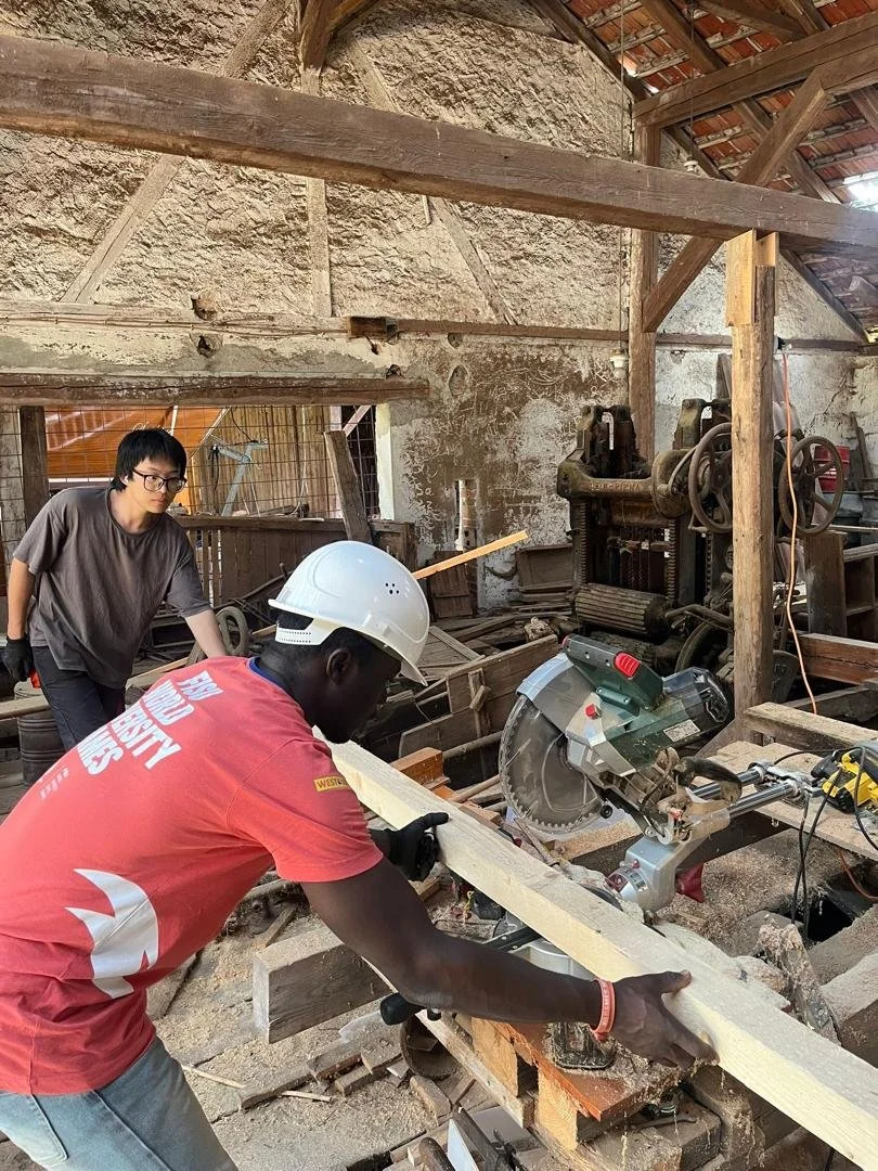 Two men working on a woodworking project in a rustic workshop; one man in a red shirt and white safety helmet is sawing a piece of wood, while the other man in a gray shirt stands nearby observing.