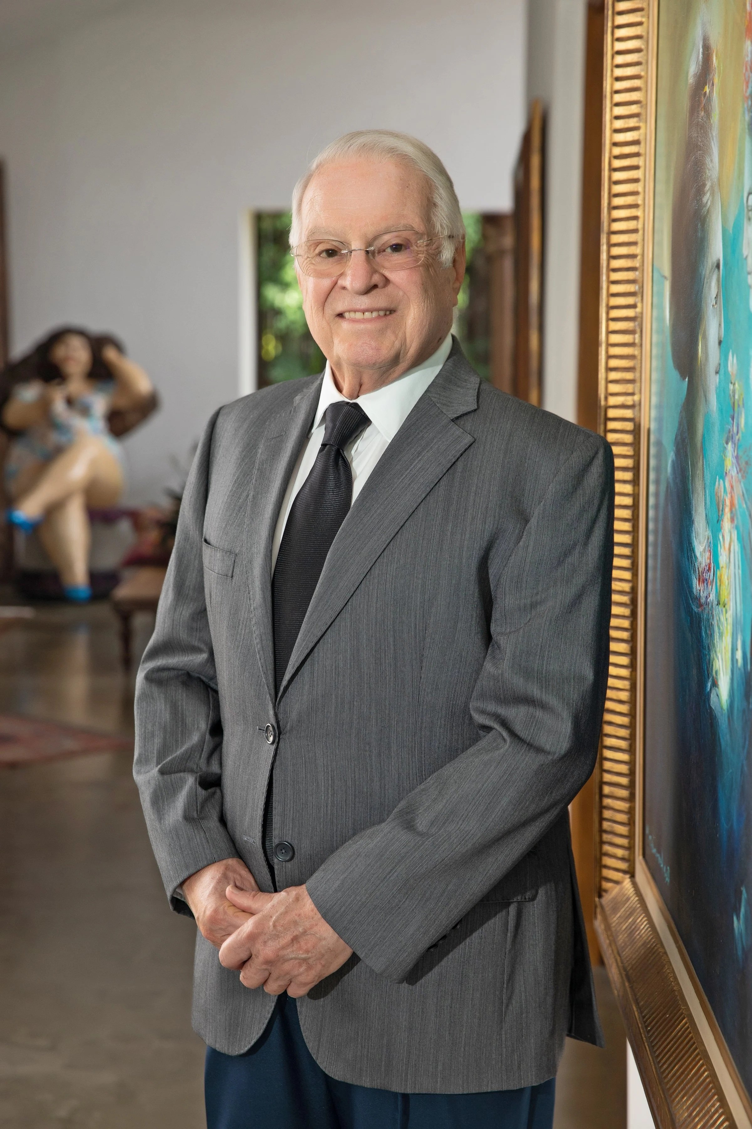 Hombre mayor con cabello canoso y gafas, vestido con saco gris y corbata negra, de pie frente a un marco con obra de arte en una galería o sala de exhibiciones.