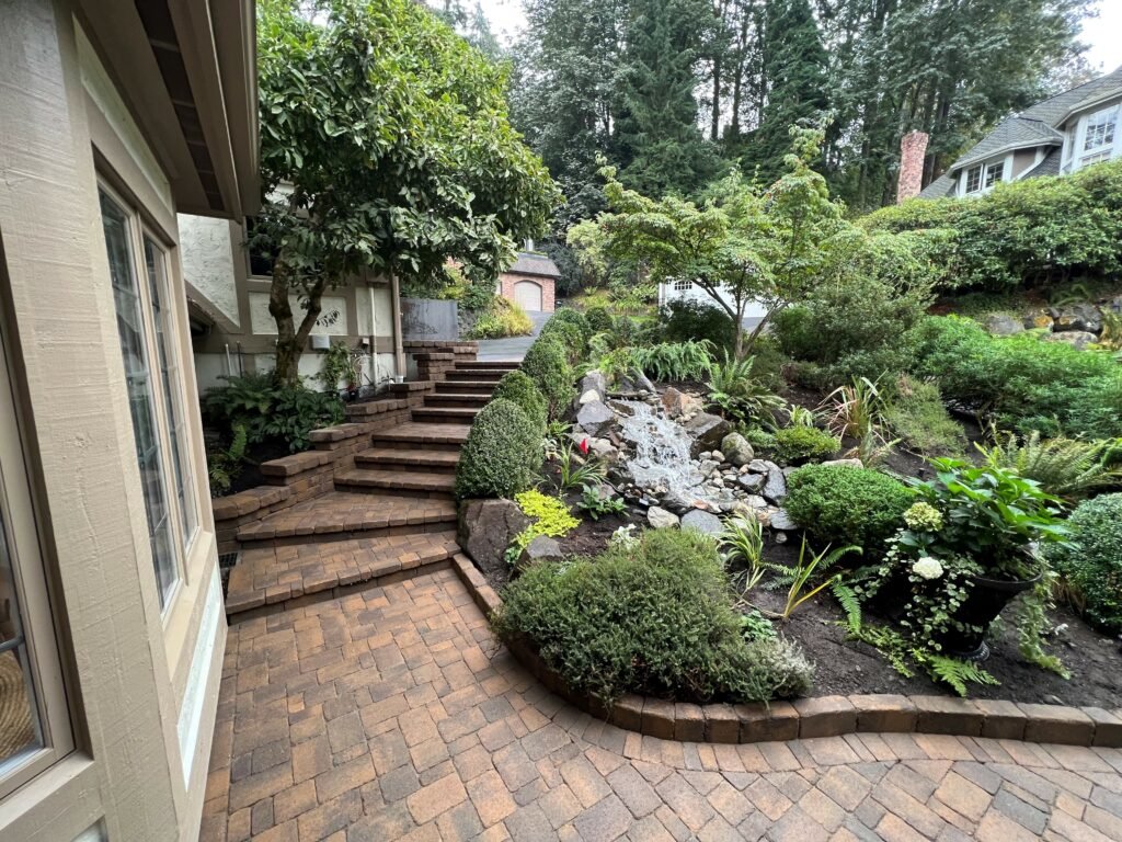 Brick patio and garden with plants, trees, and a waterfall feature in a backyard.