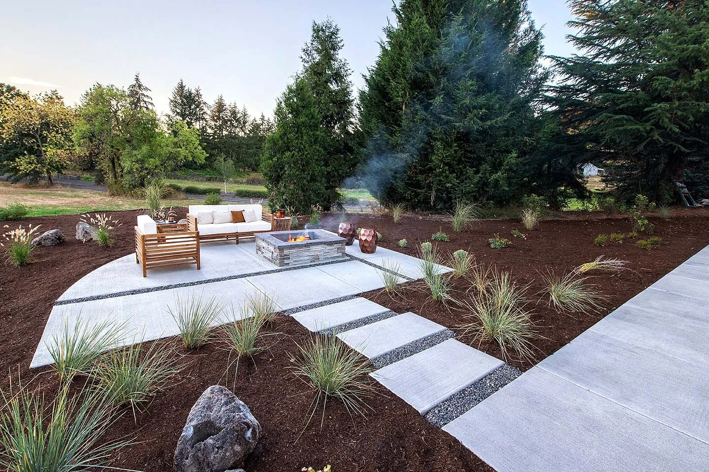Outdoor patio with a firepit, wooden and cushioned seating, potted plants, surrounded by trees and landscaped garden in the evening.