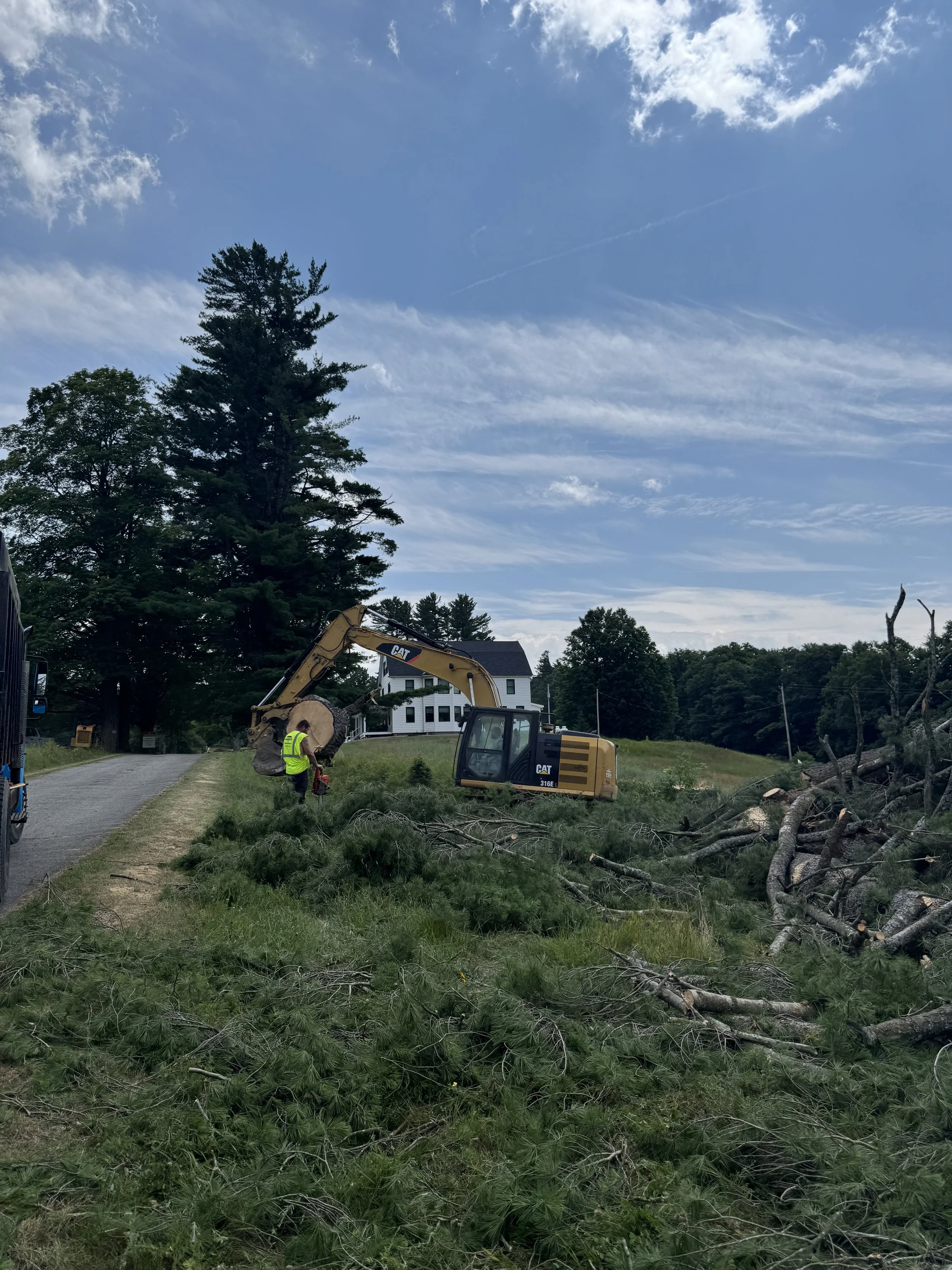 Construction scene with a yellow CAT excavator and a worker in a yellow safety vest cutting fallen tree branches on a grassy roadside under a blue sky with clouds.