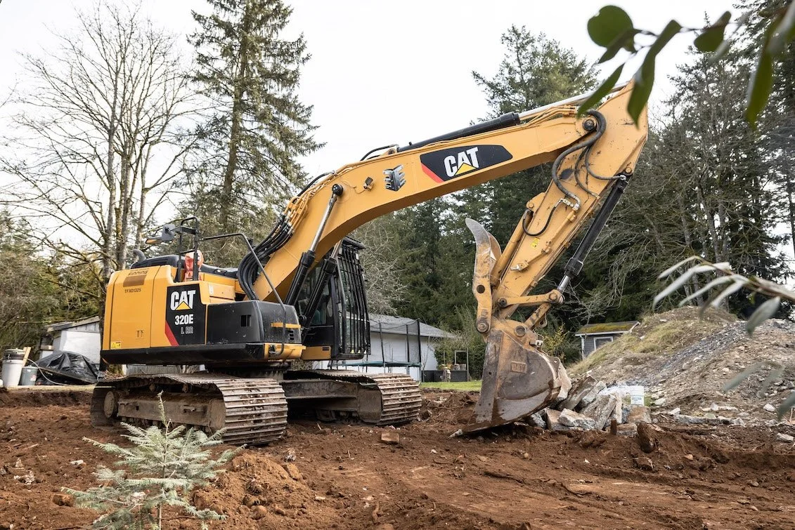 A yellow Caterpillar excavator with black tracks, labeled 320E, working outdoors on a dirt construction site with trees and small buildings in the background.