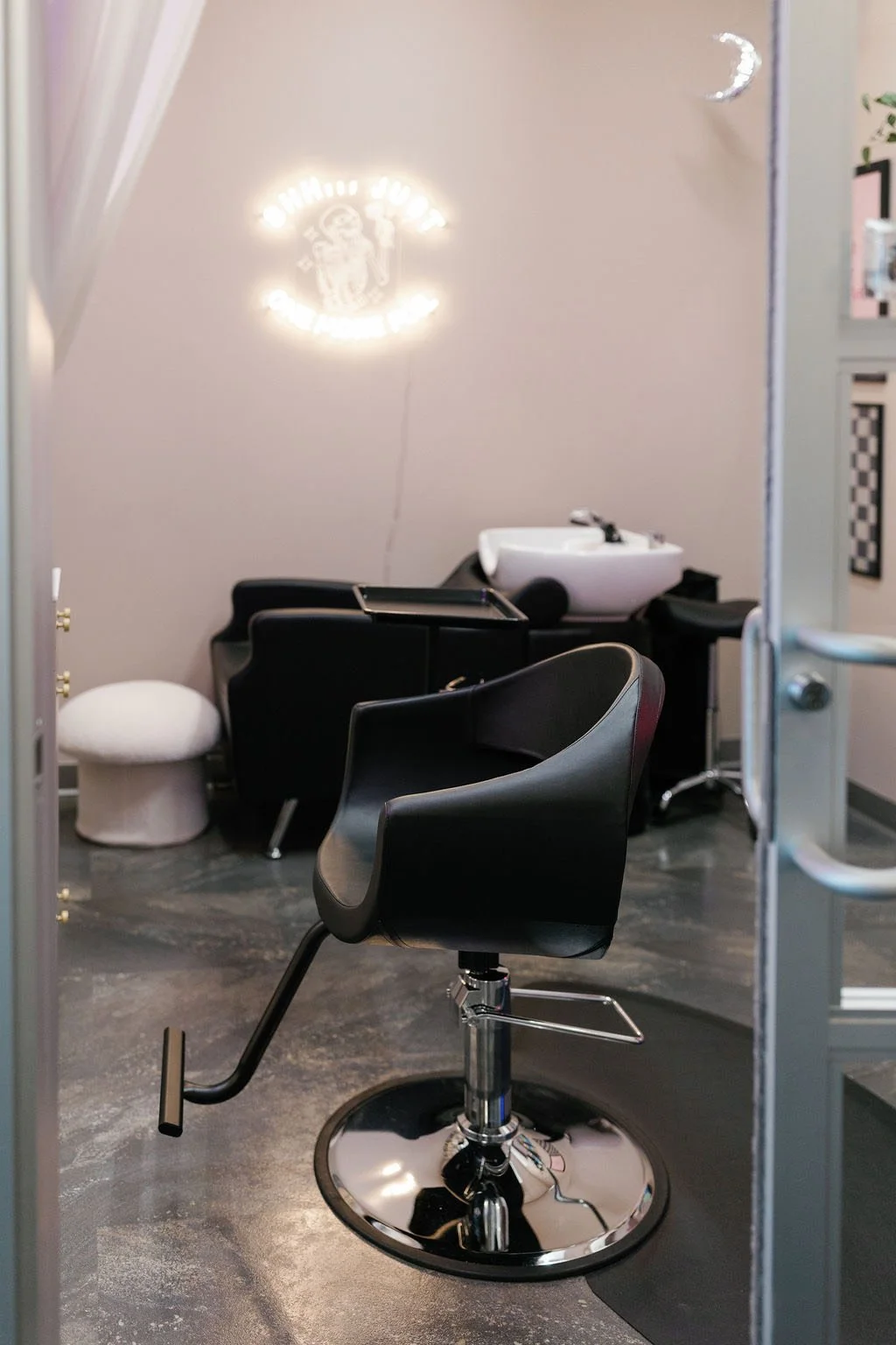 A modern hair salon  with a black styling chair on a chrome base, a small white cushioned stool, and a black sink for washing hair, with a neon sign with a skeleton on the wall reading 'JUST ONE LAST FOIL.'