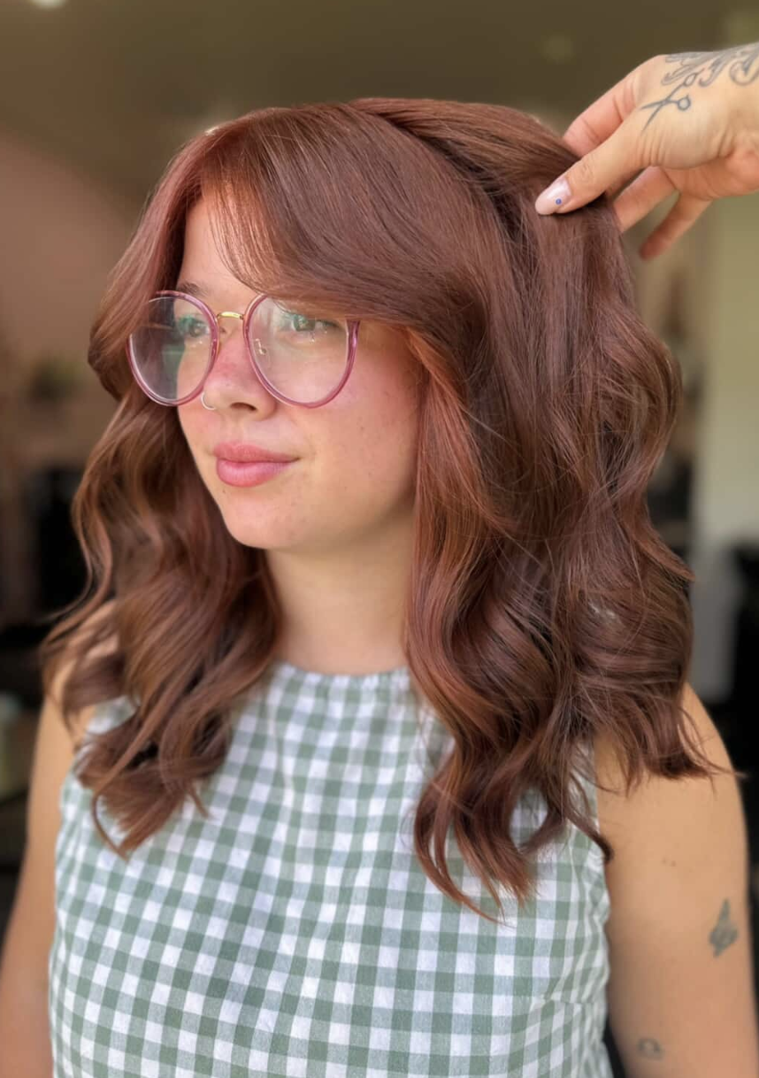 A woman with wavy, reddish-brown hair wearing pink glasses, a nose ring, and a green checkered sleeveless top.