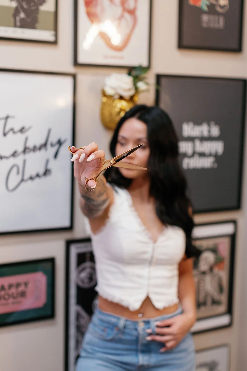 Woman with black hair and light skin holding a pair of gold scissors towards the camera in a room decorated with wall art and framed posters.