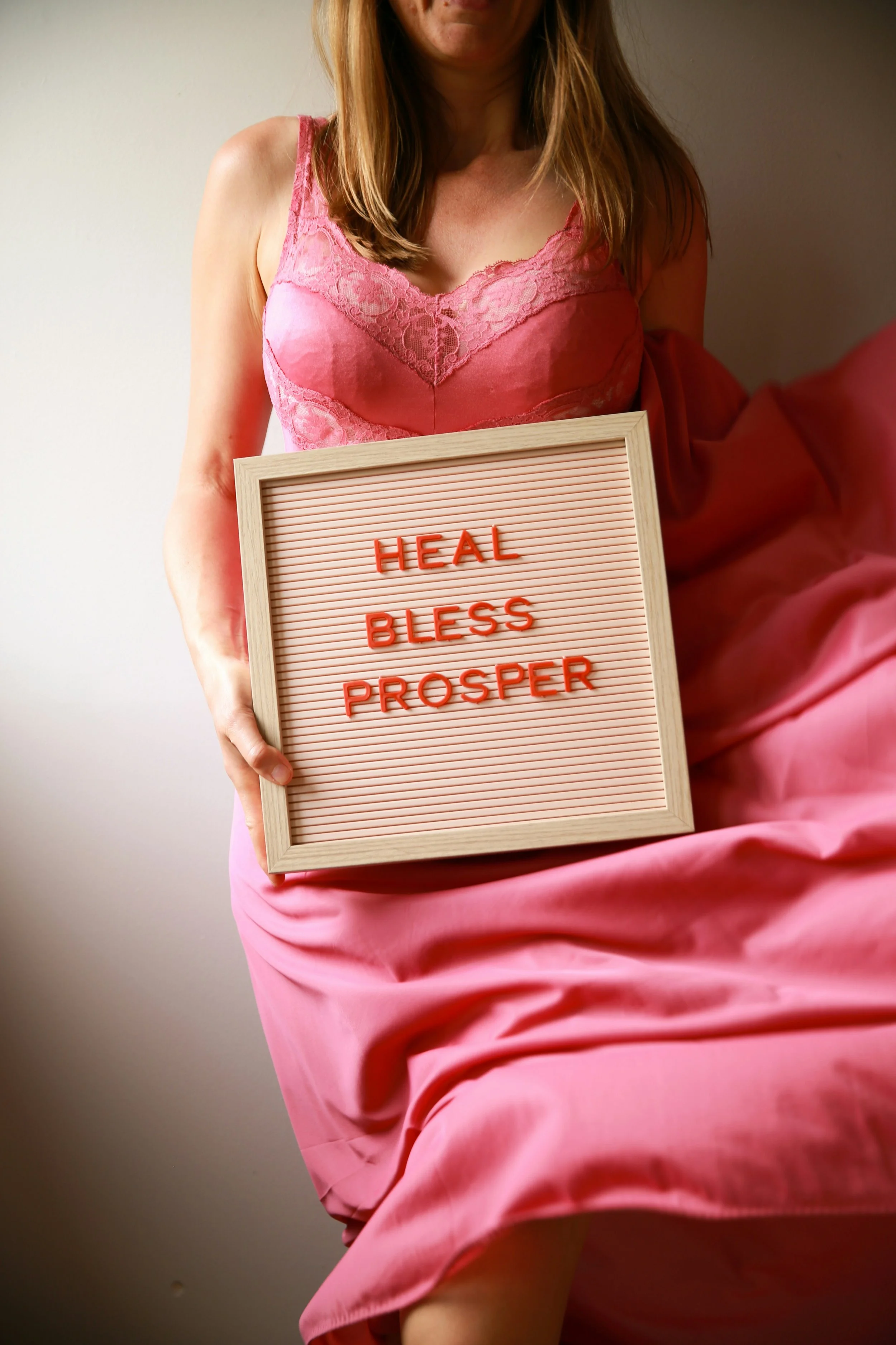 A woman dressed in pink satin lingerie holding a letter board with the message 'HEAL BLESS PROSPER' written on it. The photo is cropped to show only her torso and part of her face, with the focus on her upper body and the sign.