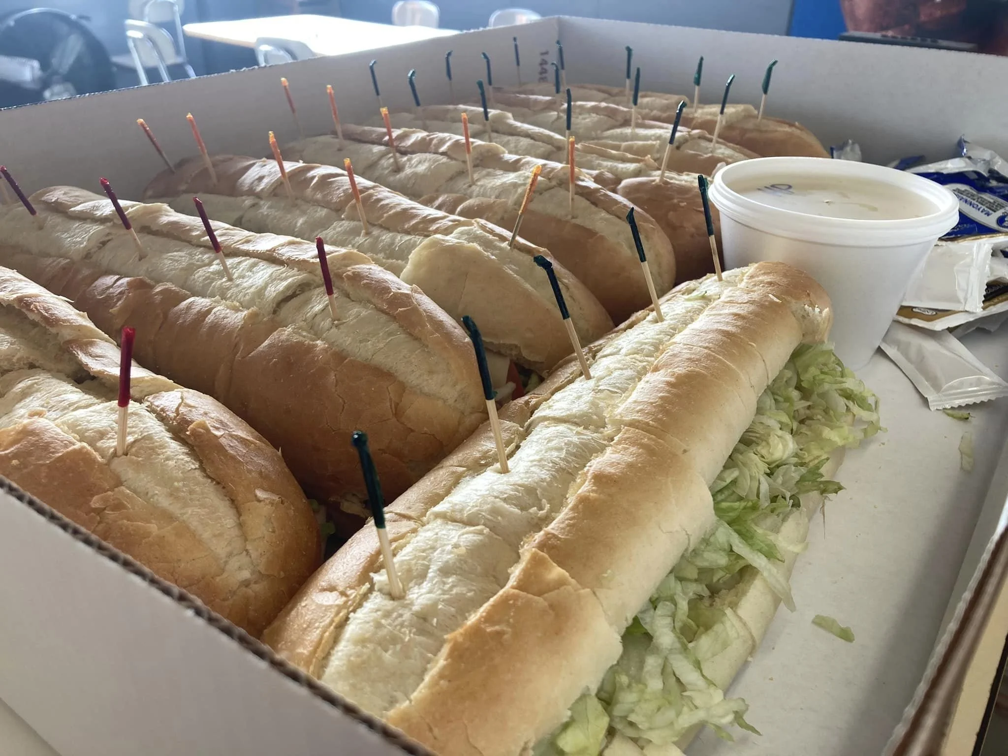 A box of assorted submarine sandwiches with shredded lettuce, accompanied by a small cup of dipping sauce.