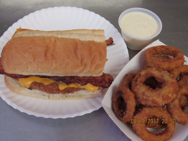 A cheeseburger sandwich with bacon on a paper plate, a side of onion rings and a cup of ranch dressing on a tray.