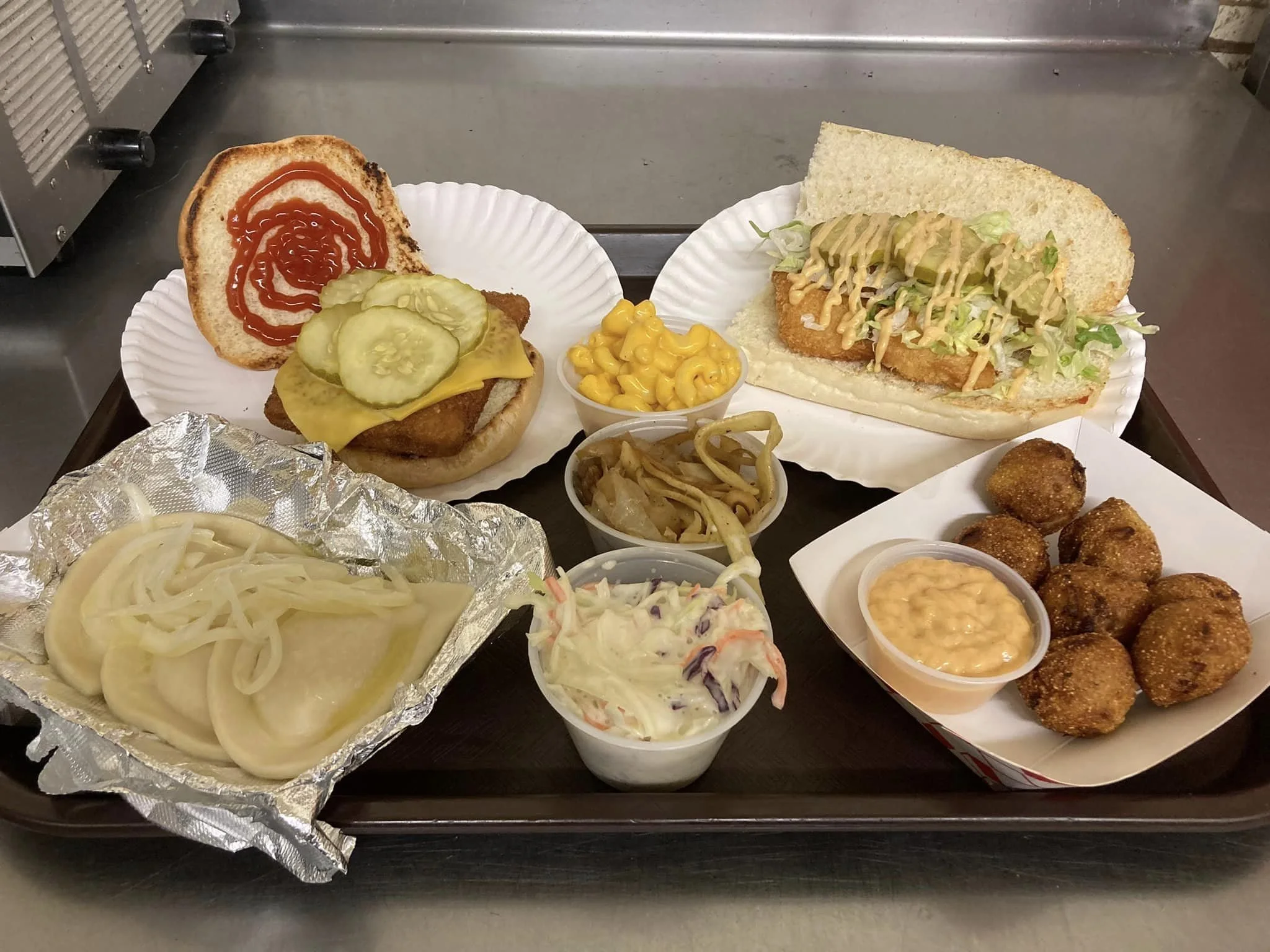 Tray of fast food items including two sandwiches, a side of macaroni and cheese, fried chicken bites with dipping sauce, onion rings, cole slaw, pickles, and onions.