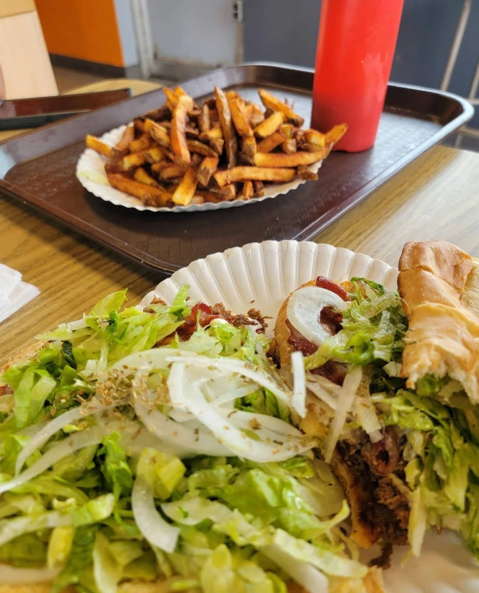 A plate of chopped lettuce, onions, and a beef burger with toppings, alongside a side of French fries and ketchup on a tray, with a sandwich in the foreground on a wooden table.