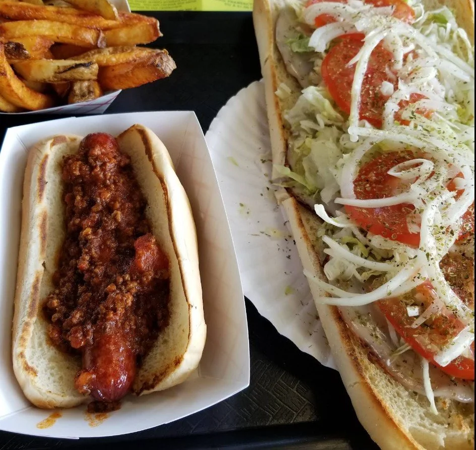 Hot dog with chili and tomato slices, French fries, and a sub sandwich with lettuce, tomatoes, and onions on a sandwich bun.