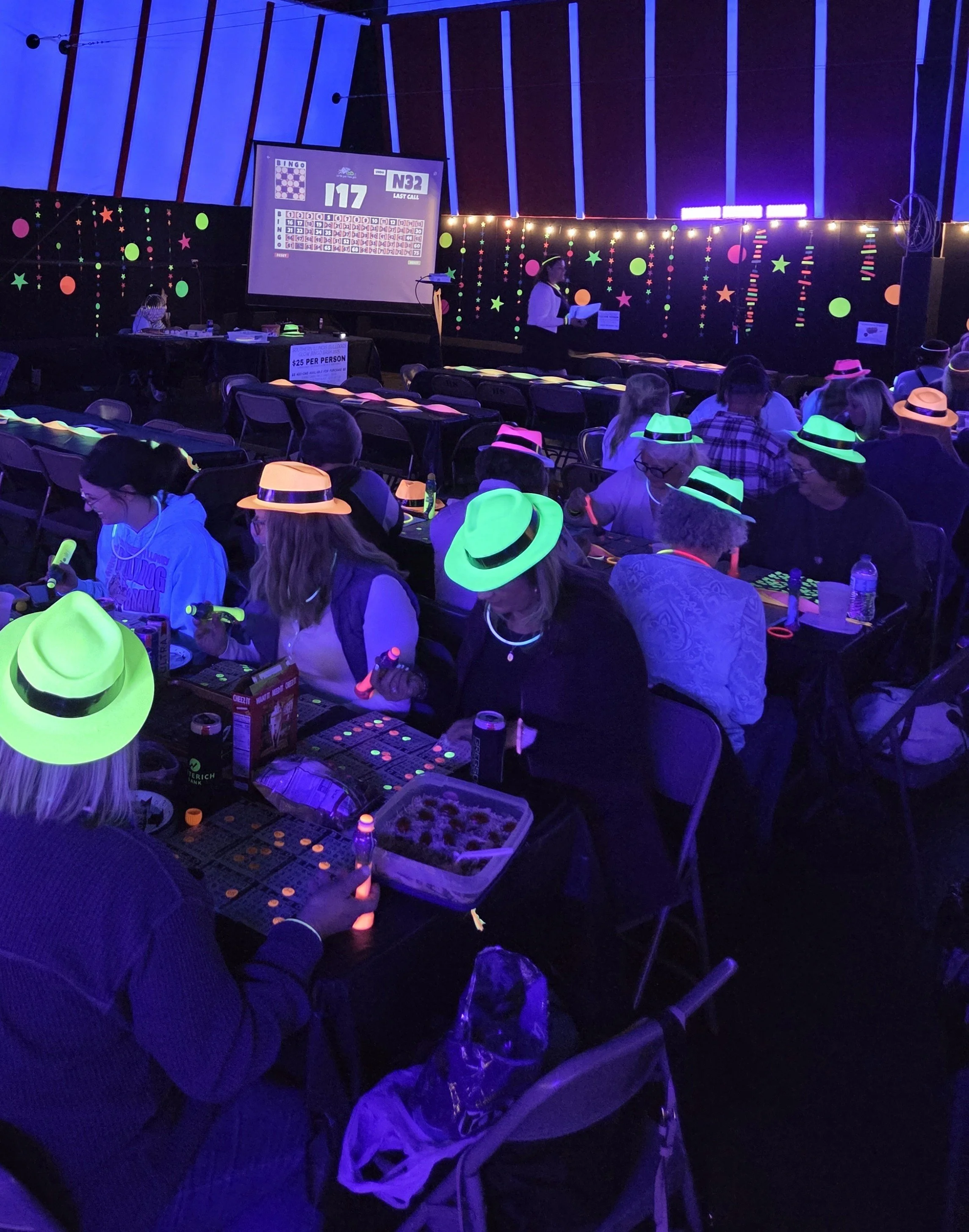 People wearing neon hats participating in a glow-in-the-dark bingo game in a dimly lit room decorated with colorful glowing stars and dots, with a large screen displaying bingo numbers.