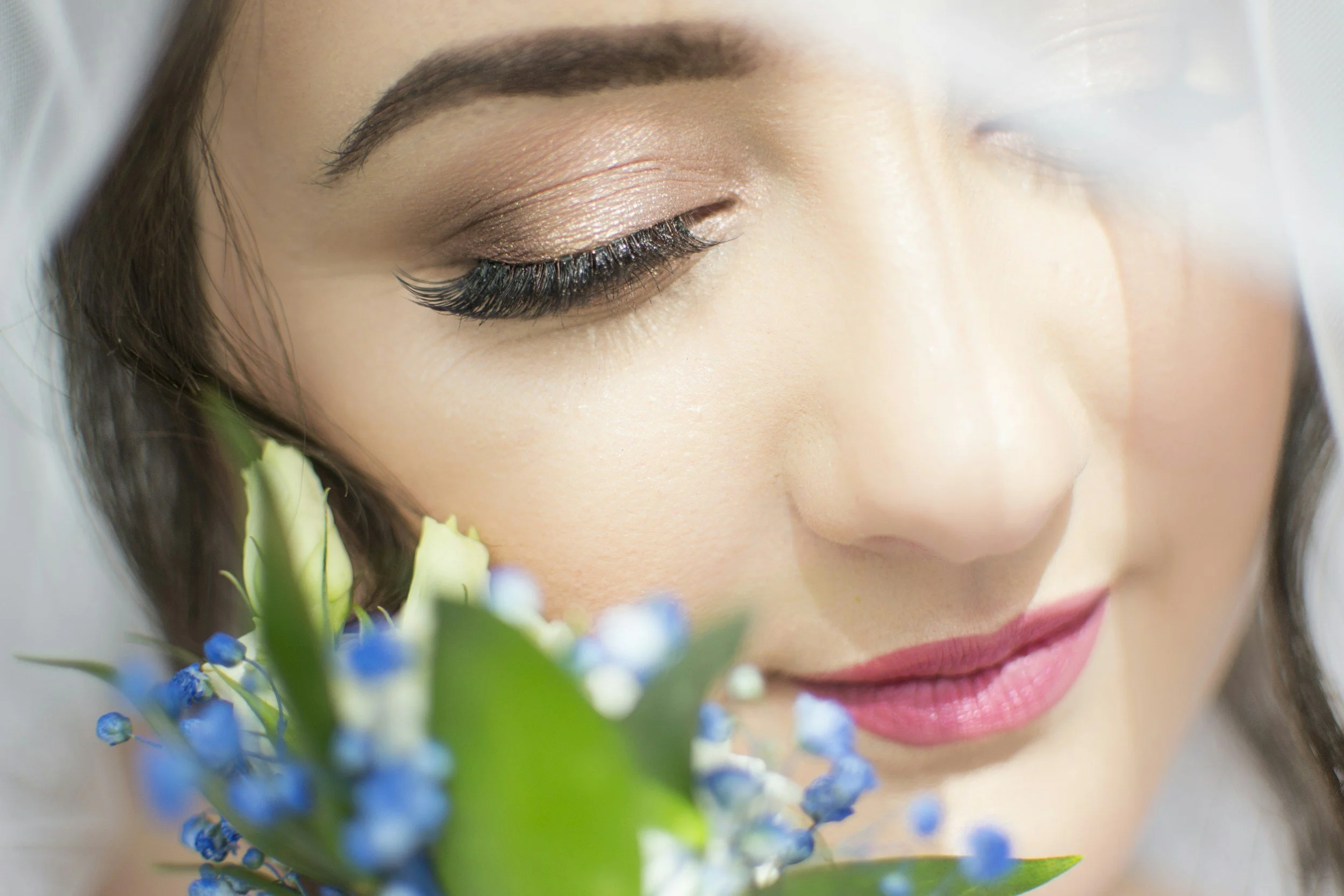 A close-up of a woman with makeup and long eyelashes holding a bouquet of small blue flowers with green leaves.