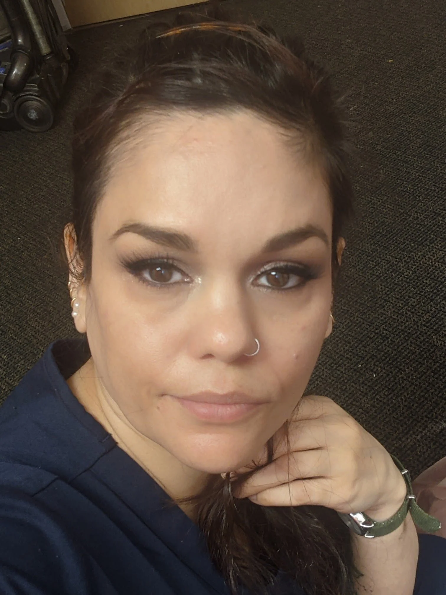 A woman with brown hair and makeup, wearing a dark shirt, sitting on a carpeted floor with a stroller in the background.
