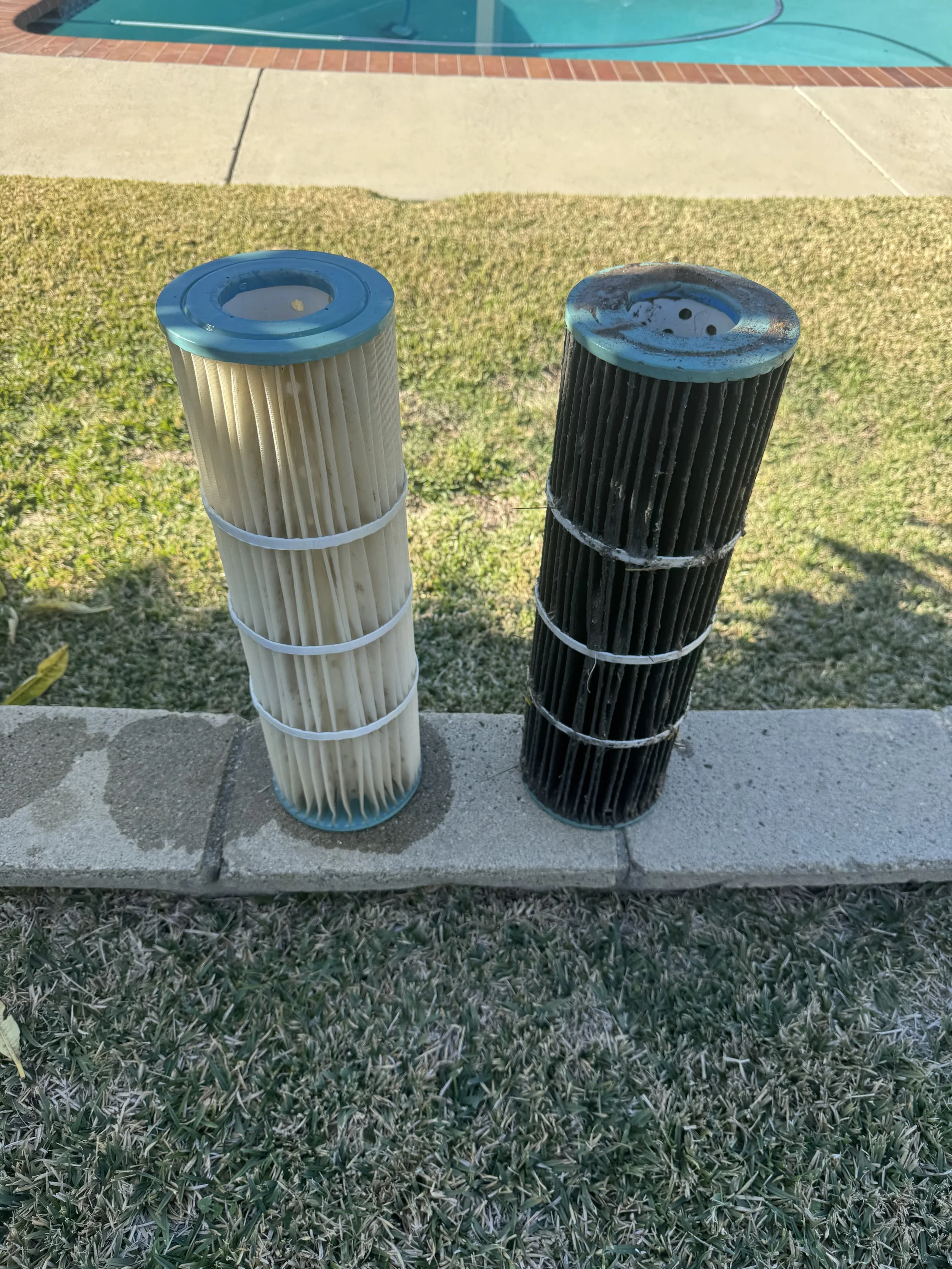 Two pool filters, one clean and white, the other dirty and black, placed on a low concrete wall near a lawn and swimming pool.