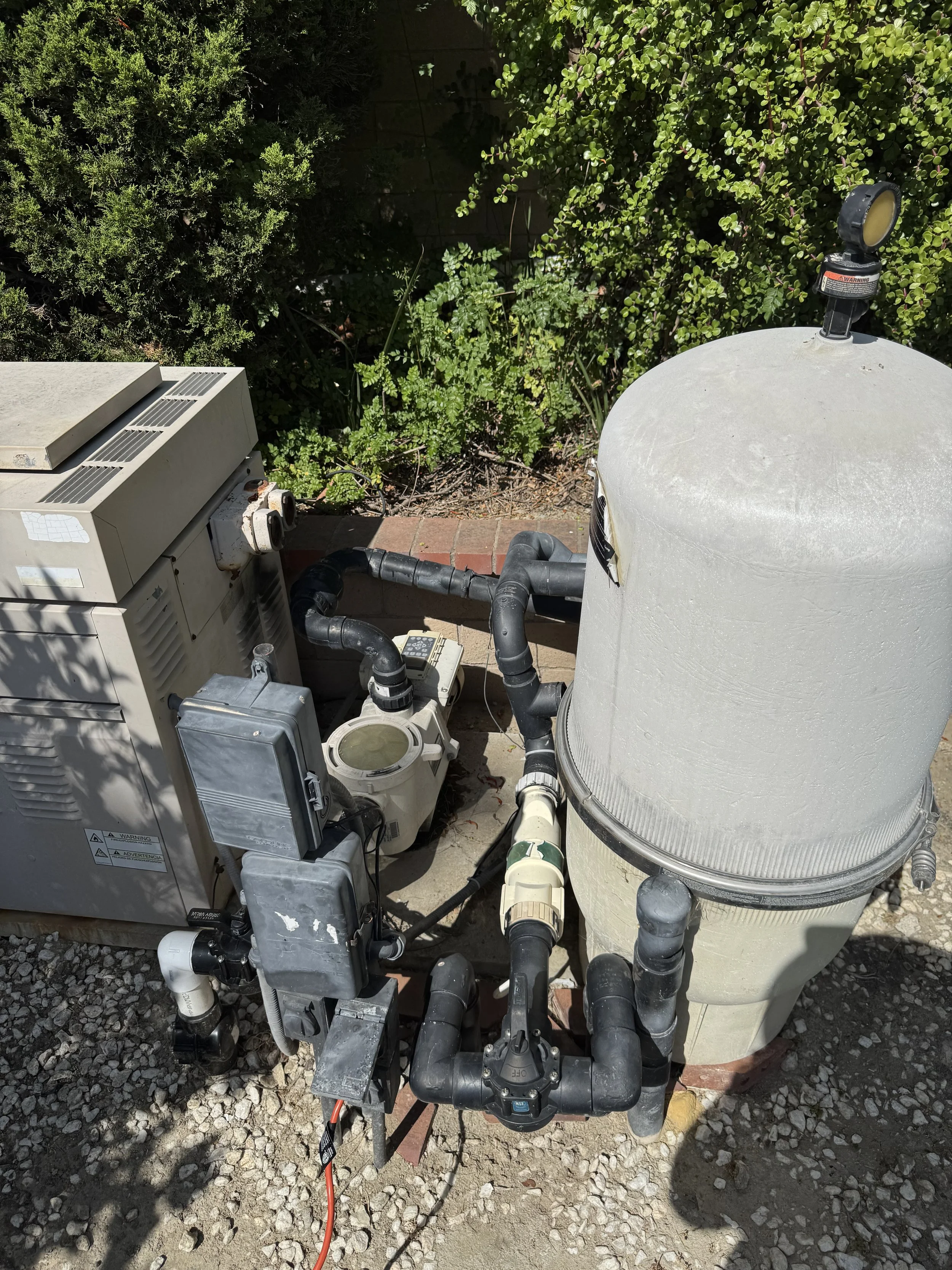 Pool pump system with pipes, filter and motor next to green bushes on gravel ground.