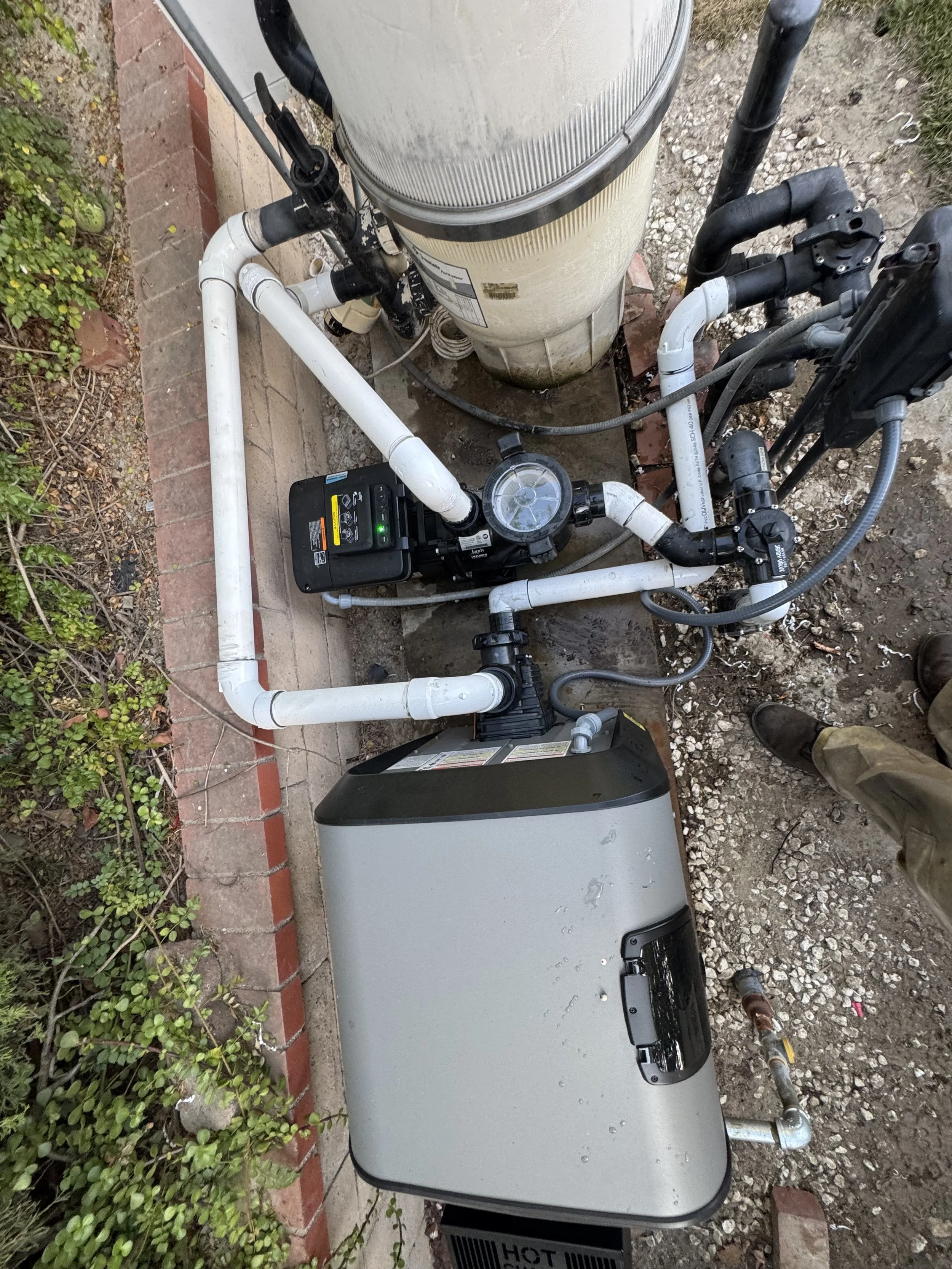 Pool pump system with pipes, filter, and control box next to a landscaped area with bricks.