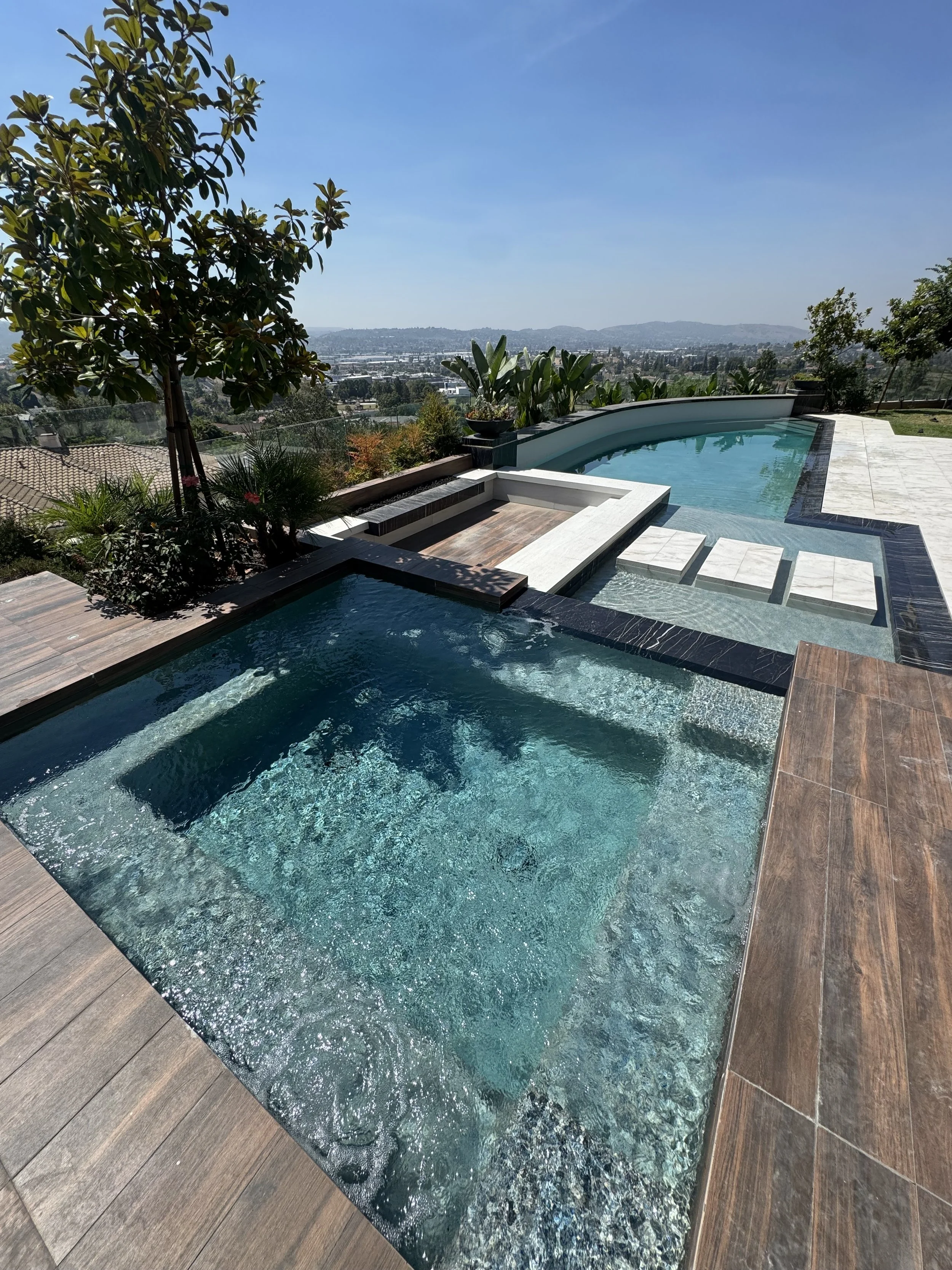 Luxury outdoor pool area with a hot tub and a naturalistic landscape view, including trees and a cityscape in the distance.