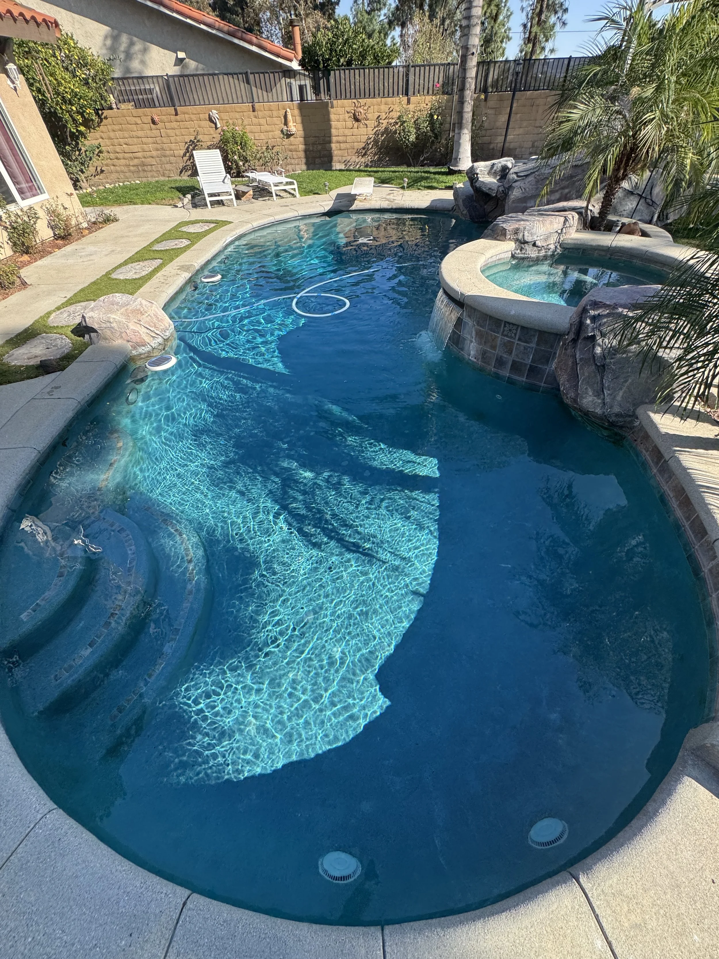 Swimming pool with steps, hot tub, rocks, palm tree, lounge chairs, and backyard with fence.