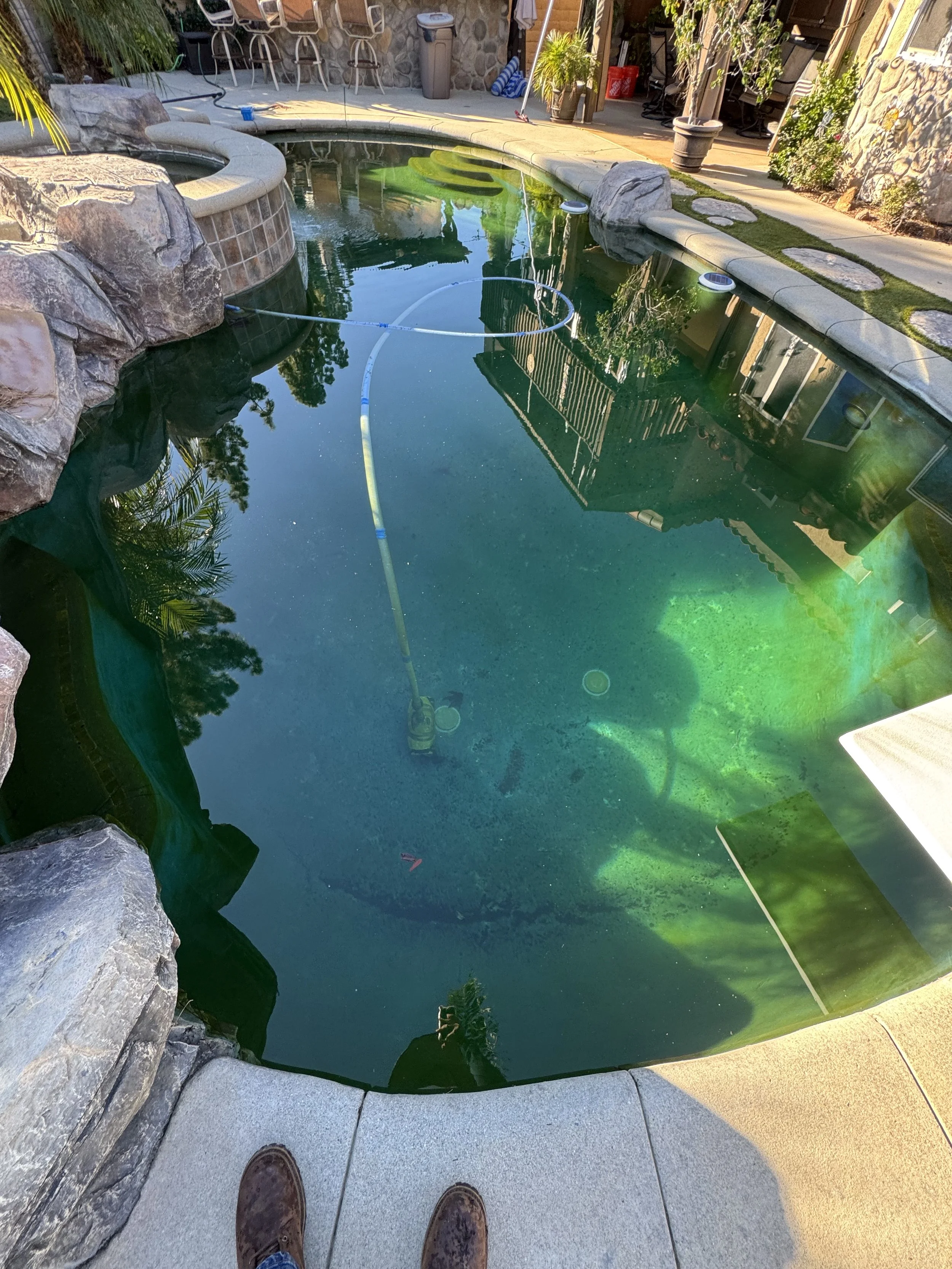 Empty swimming pool with greenish water, surrounded by rocks and patio furniture, with a person’s feet visible at the bottom of the image.