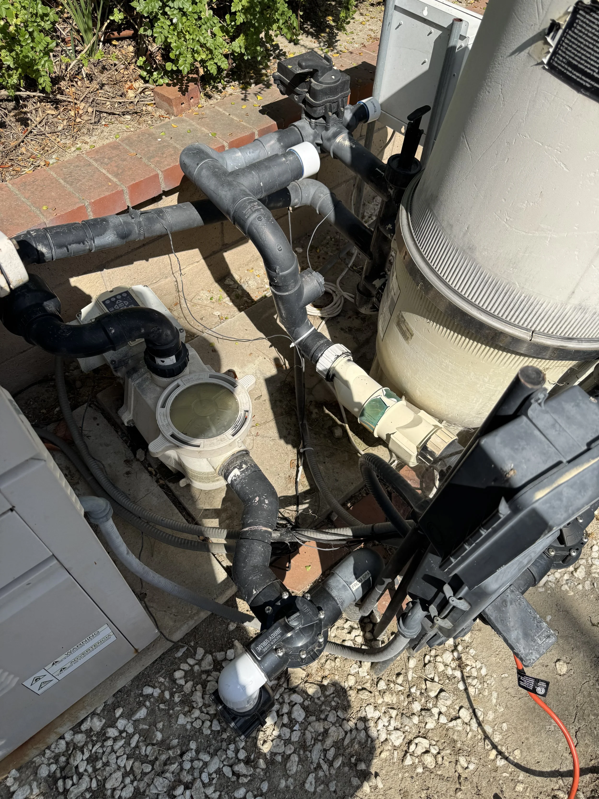 A pool pump system with black pipes, a white pool filter, and an electrical control box. The setup is outdoors on a concrete surface near a garden bed with green plants and a brick border.