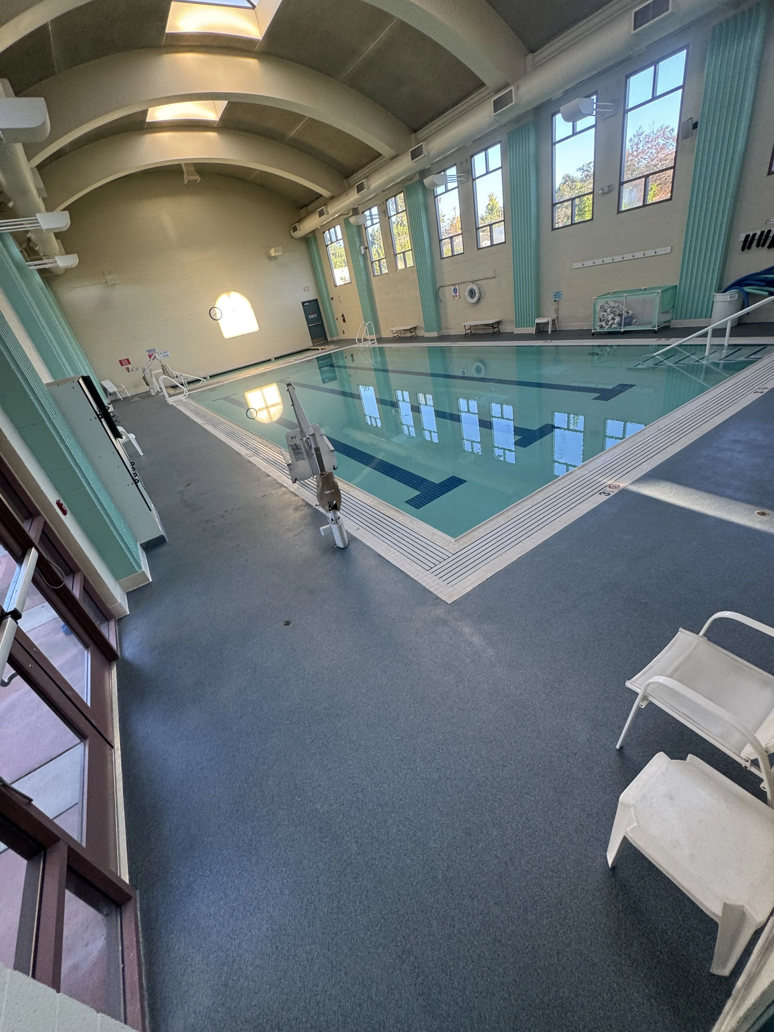 Indoor swimming pool with large windows, empty pool, surrounded by a grey non-slip floor, with white and aqua walls, chairs, and pool equipment.