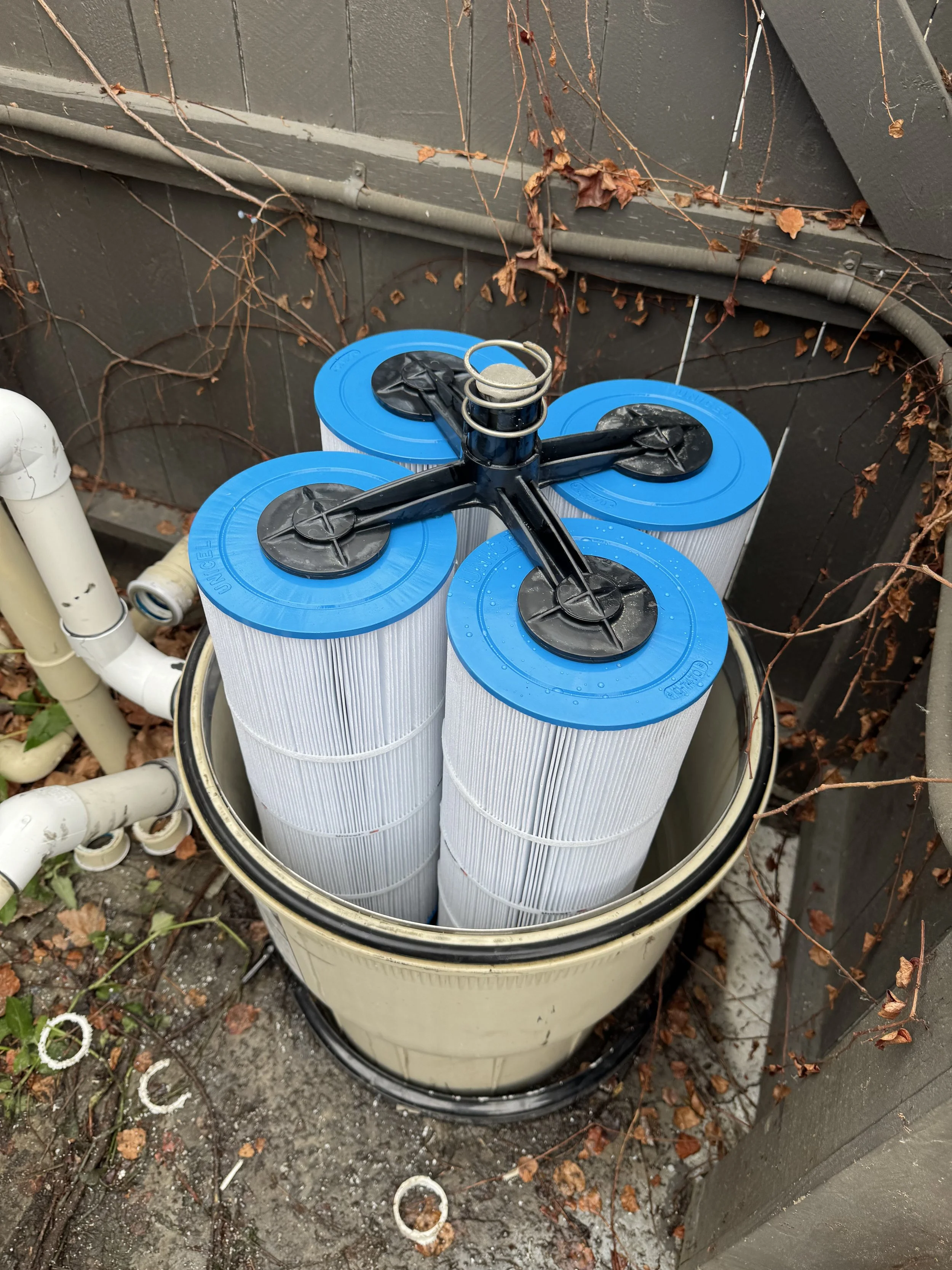 A water filter system with four large white filter cartridges and a black plastic wrench on top, placed inside a beige bucket outdoors next to a gray wall with dried vines.
